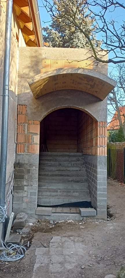 Budowa wejścia do domu z cegły i betonu, widoczne schody i łukowe zadaszenie w surowym stanie, teren wokół jeszcze niezagospodarowany.