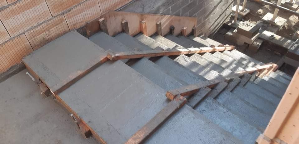 Wylewanie betonowych schodów wewnętrznych z drewnianym szalunkiem i ceglaną ścianą w tle.