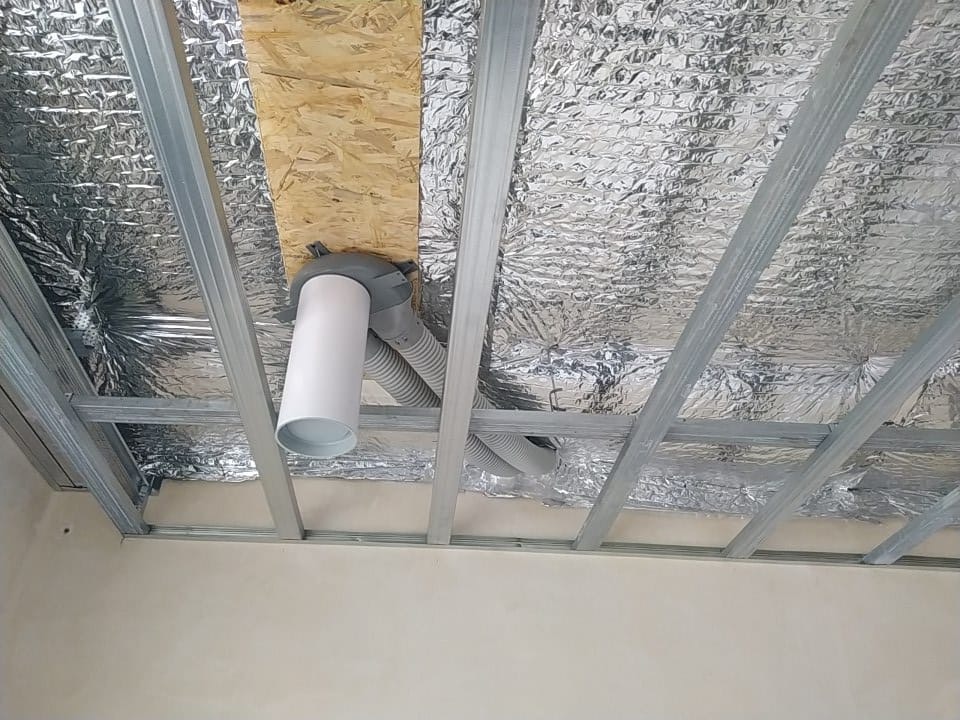 Instalacja kanałów wentylacyjnych pod sufitem podwieszanym z folią izolacyjną, widoczne rury i metalowa konstrukcja nośna.