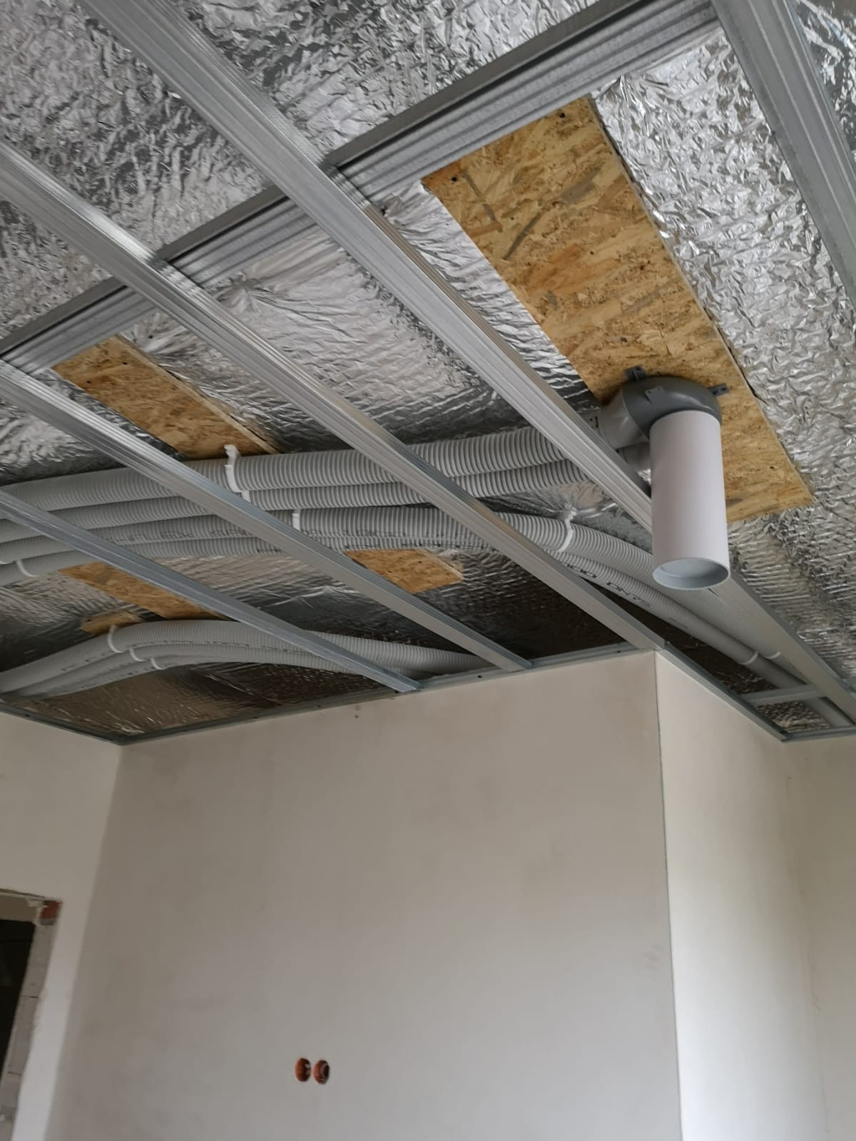 Instalacja przewodów wentylacyjnych w przestrzeni pod sufitem podwieszanym, widoczne rury karbowane, izolacja folią aluminiową i elementy mocujące do płyty OSB.