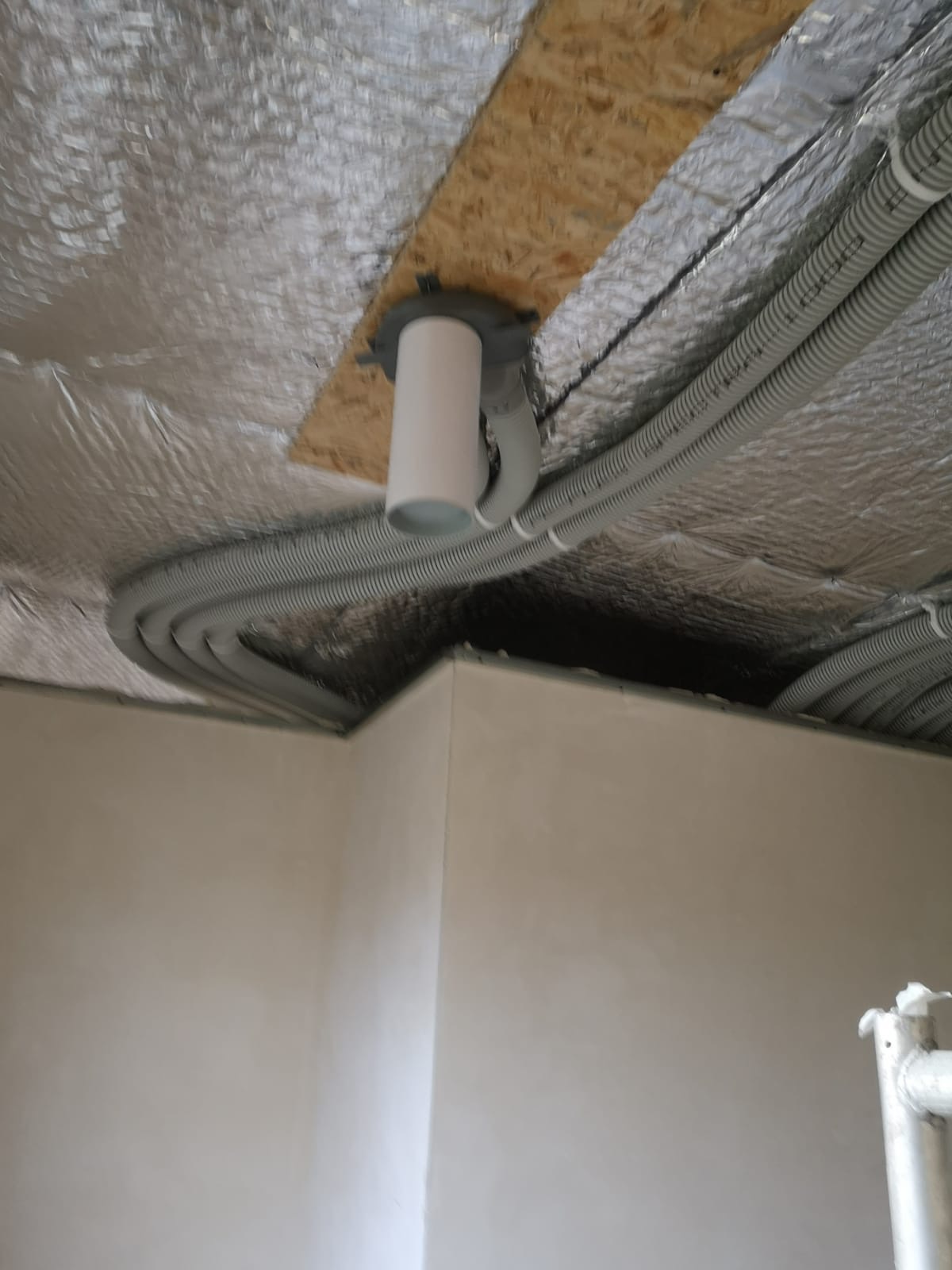Szare, elastyczne rury wentylacyjne zamontowane pod folią izolacyjną na suficie, połączone z białym anemostatem, widok z dołu na róg pomieszczenia ze ścianami w kolorze złamanej bieli.