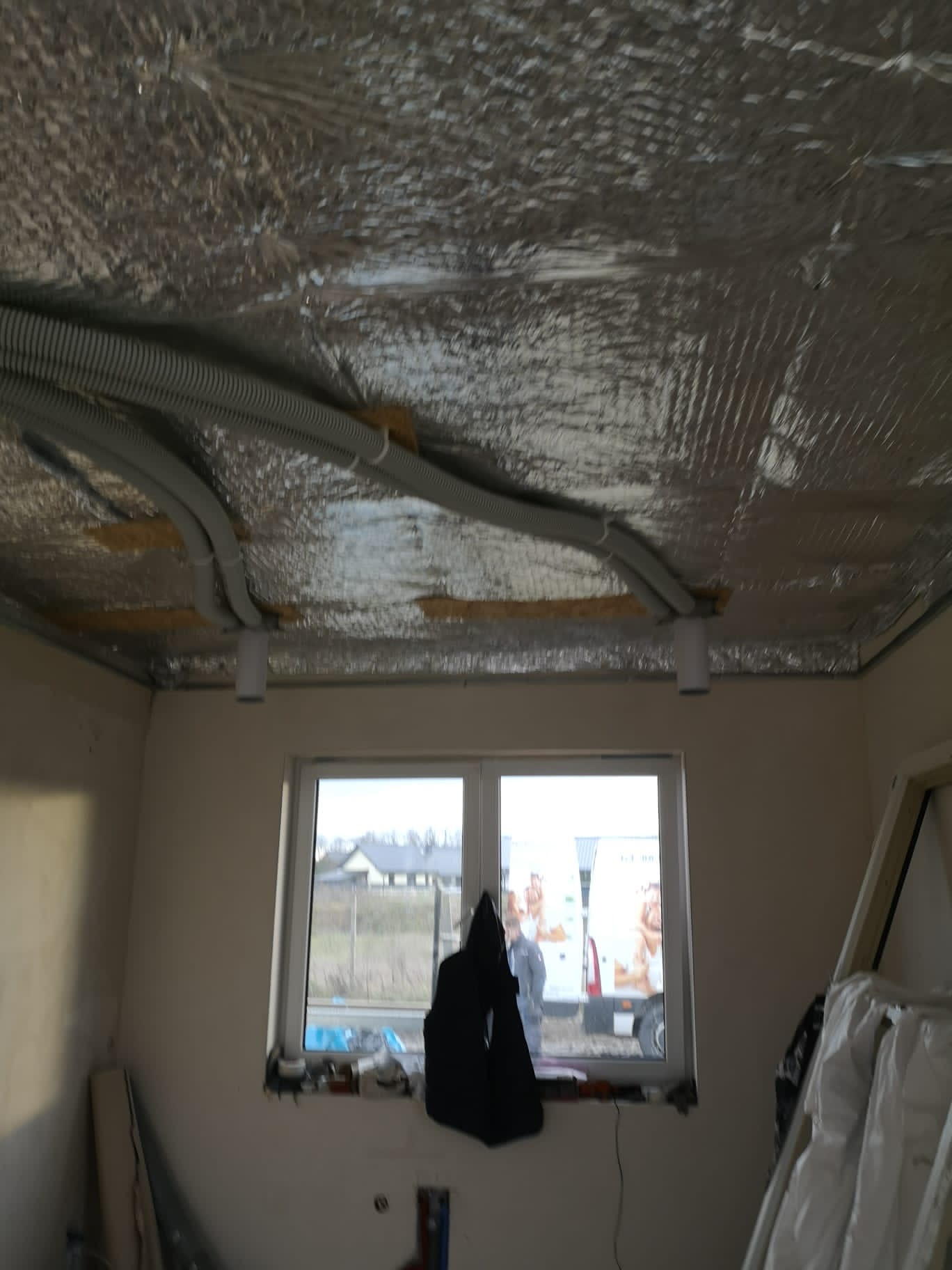 Wnętrze w trakcie prac wykończeniowych z widocznymi przewodami wentylacyjnymi pod folią izolacyjną na suficie, okno z widokiem na zabudowę jednorodzinną i zaparkowany samochód dostawczy.