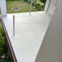 Wykończony taras z szarych płytek ceramicznych, widok z góry na ogród z miejscem na ognisko, metalowe słupki balustrady, fragment ściany budynku w prawym dolnym rogu.