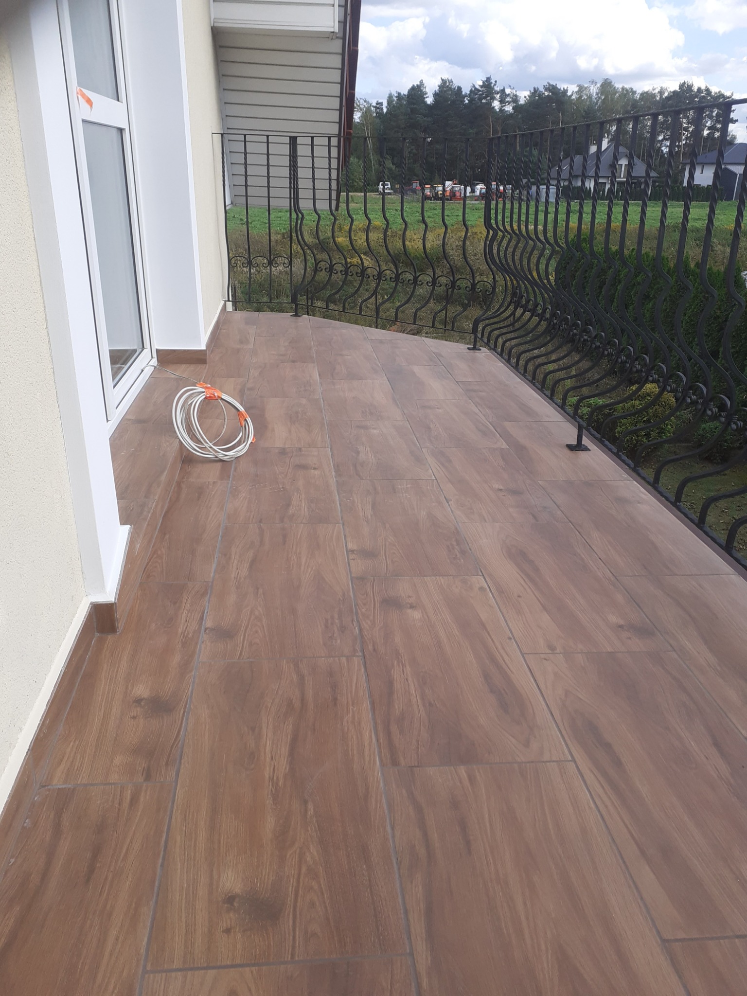 Wykończony balkon z brązowymi płytkami imitującymi drewno, czarna, ozdobna balustrada i widok na zielony ogród z domami w tle; na podłodze zwój białego kabla.