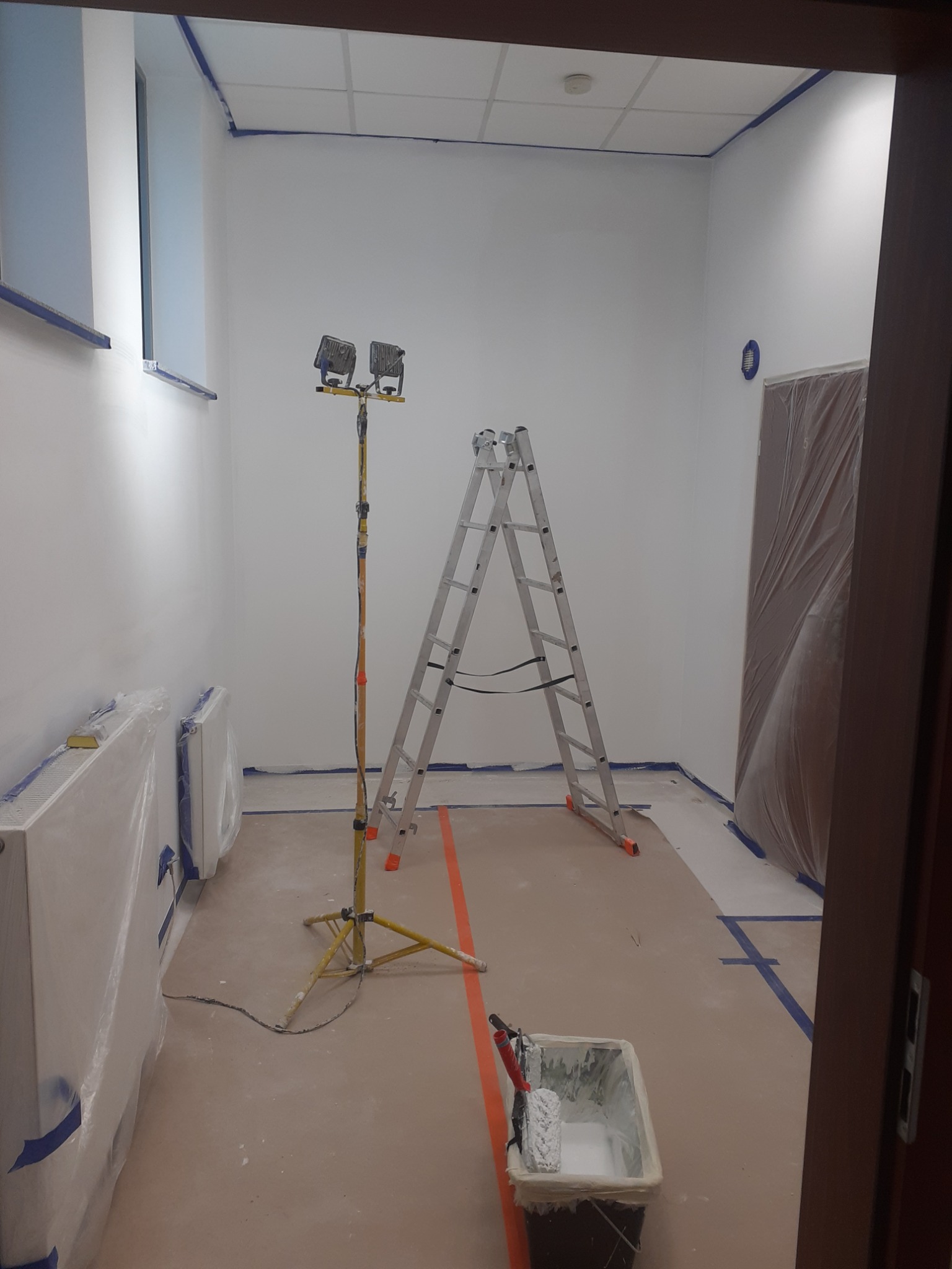 Pokój przygotowany do malowania: ściany zabezpieczone taśmą malarską, grzejniki i drzwi osłonięte folią, na podłodze wiadro z farbą i wałek, drabina aluminiowa oraz lampa halogenowa na statywie.