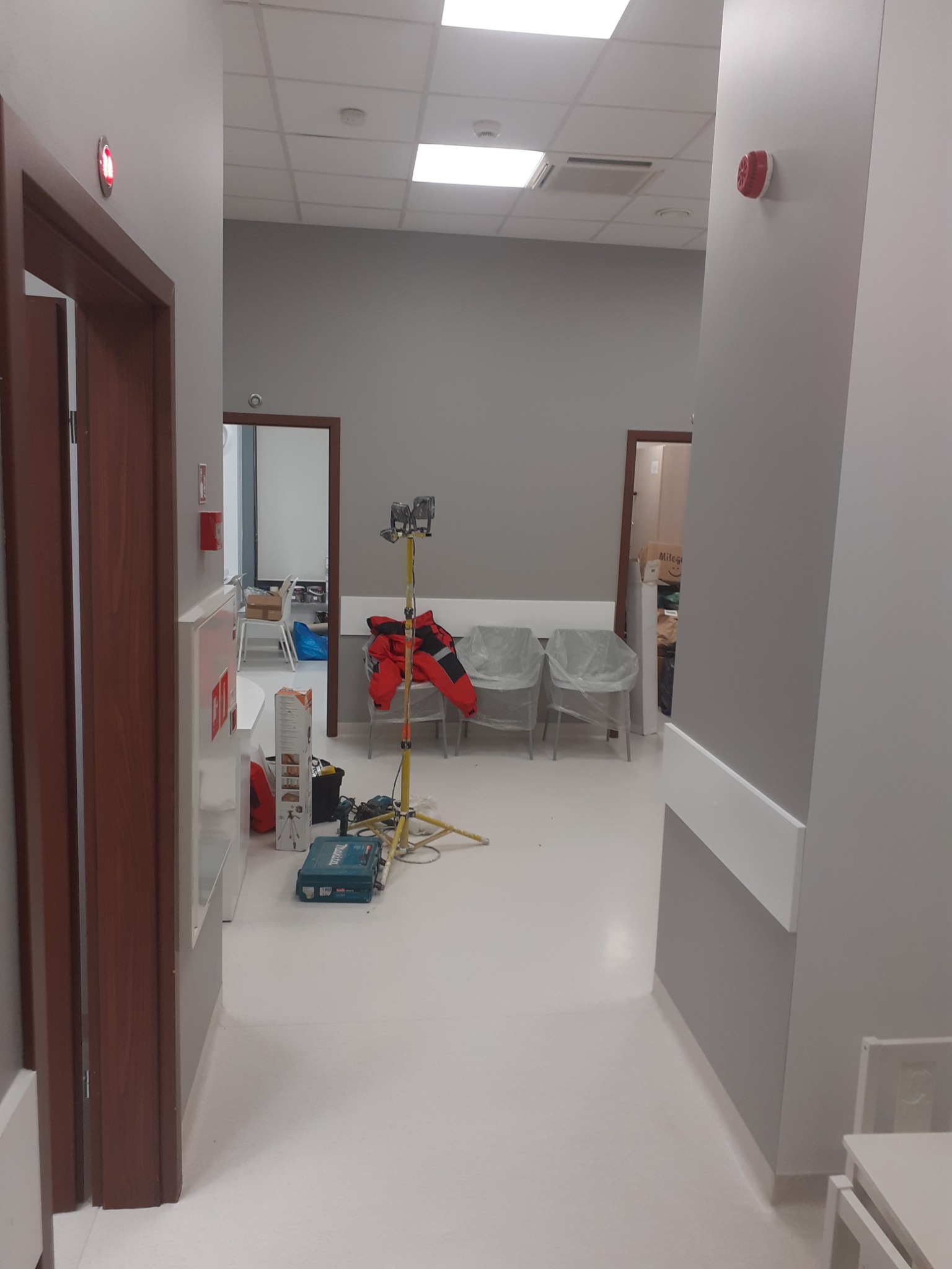 Korytarz w trakcie malowania, widoczny sprzęt malarski, lampa oświetleniowa i narzędzia, ściany w odcieniach szarości, biała podłoga.
