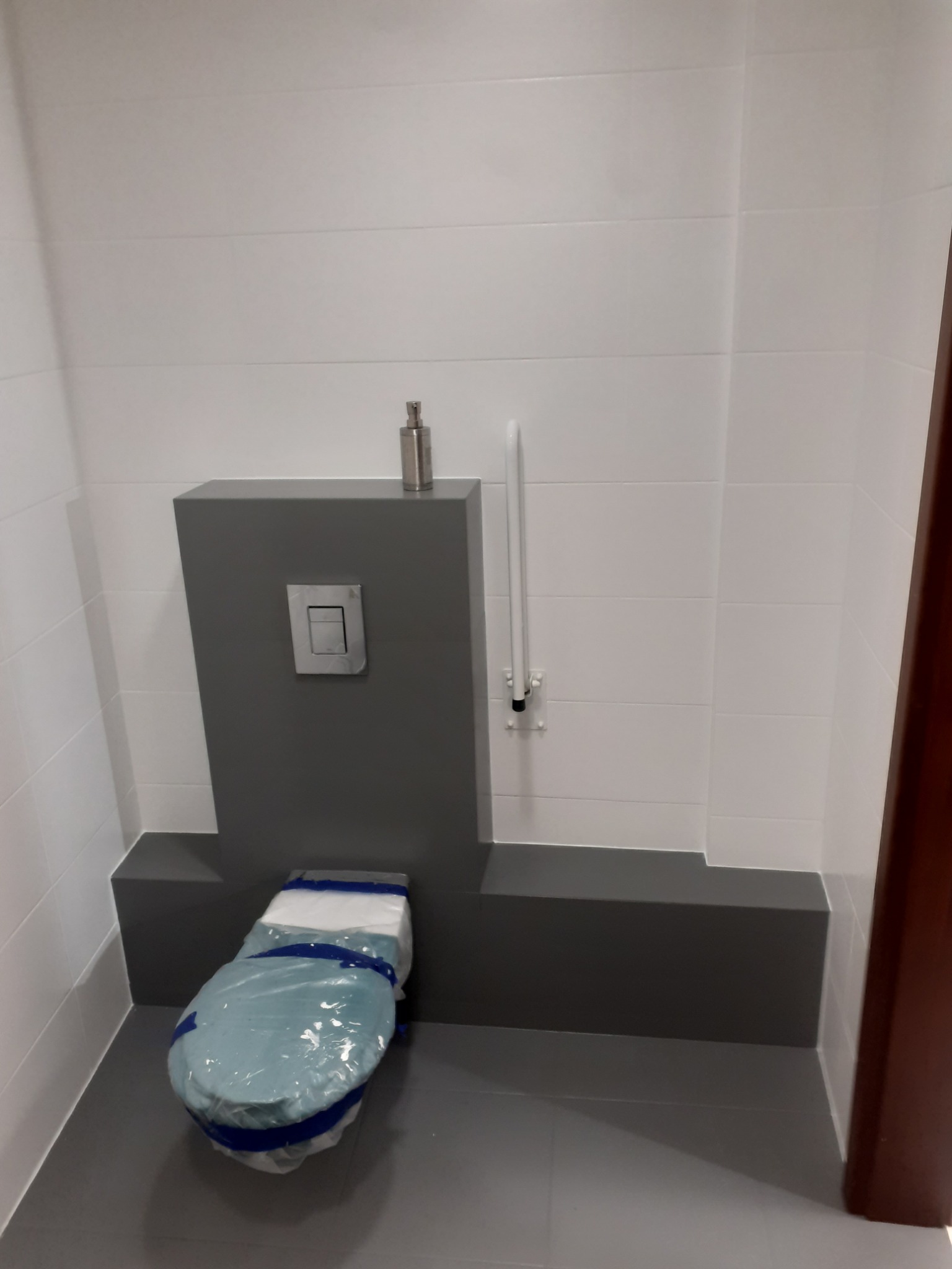 Wisząca toaleta w trakcie montażu, zabezpieczona folią ochronną, z widoczną spłuczką podtynkową i uchwytem dla niepełnosprawnych na białych płytkach.