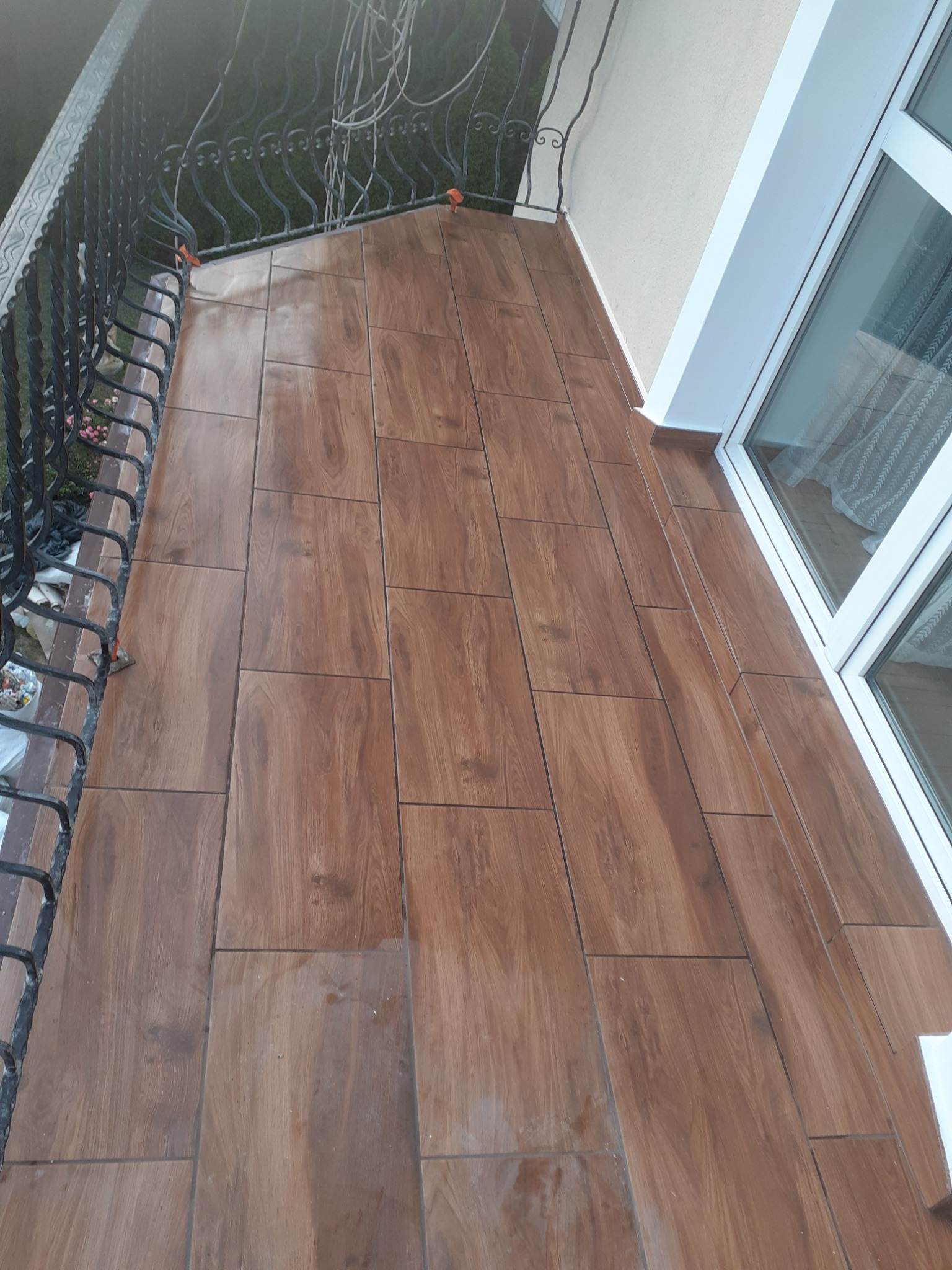 Balkon z nową podłogą wyłożoną brązowymi płytkami imitującymi drewno, widoczna barierka z kutego żelaza i okno balkonowe.