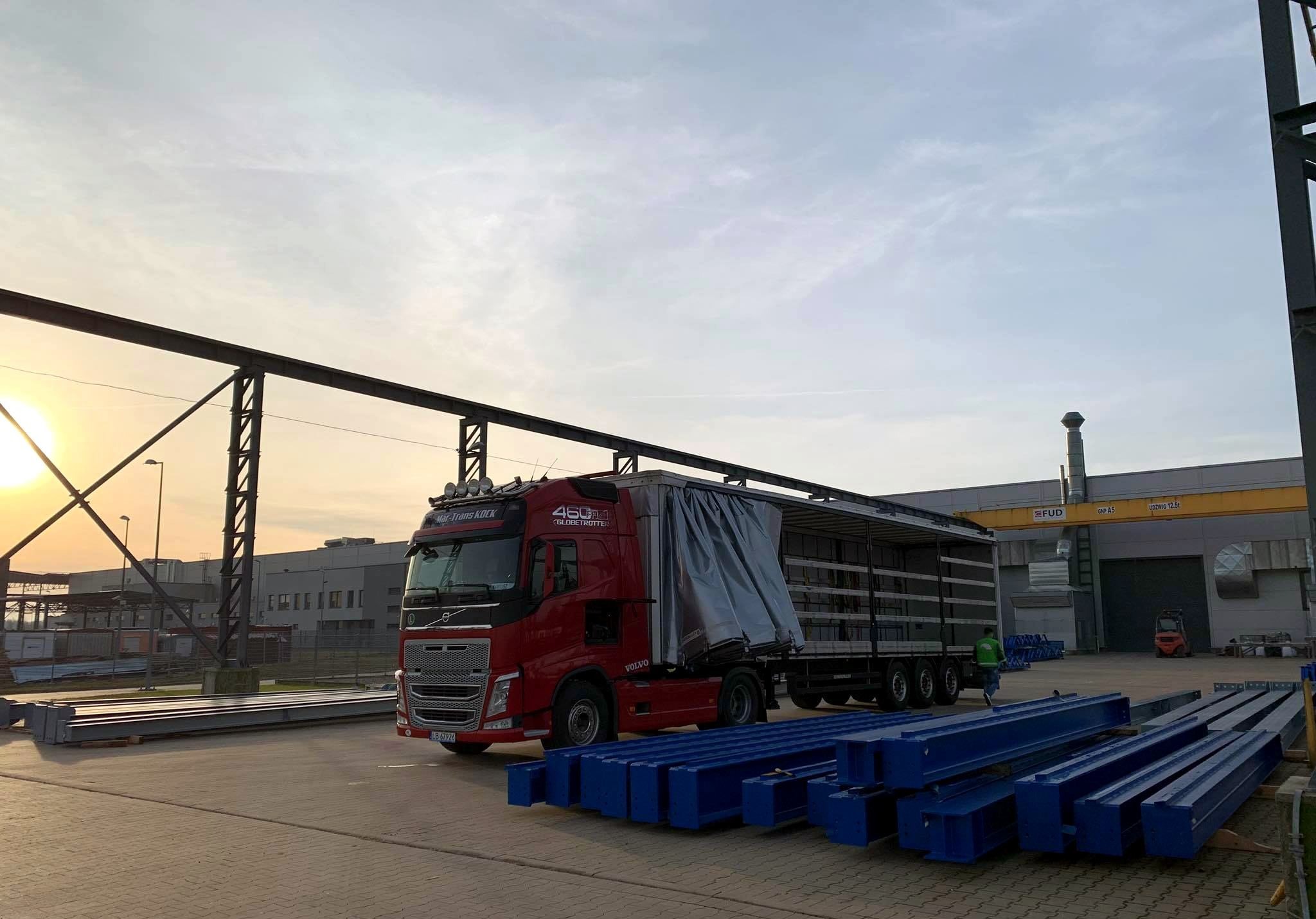 Czerwona ciężarówka Volvo z naczepą kurtynową na placu budowy, obok leżą pomalowane na niebiesko stalowe elementy konstrukcyjne, w tle budynek przemysłowy i zachodzące słońce.