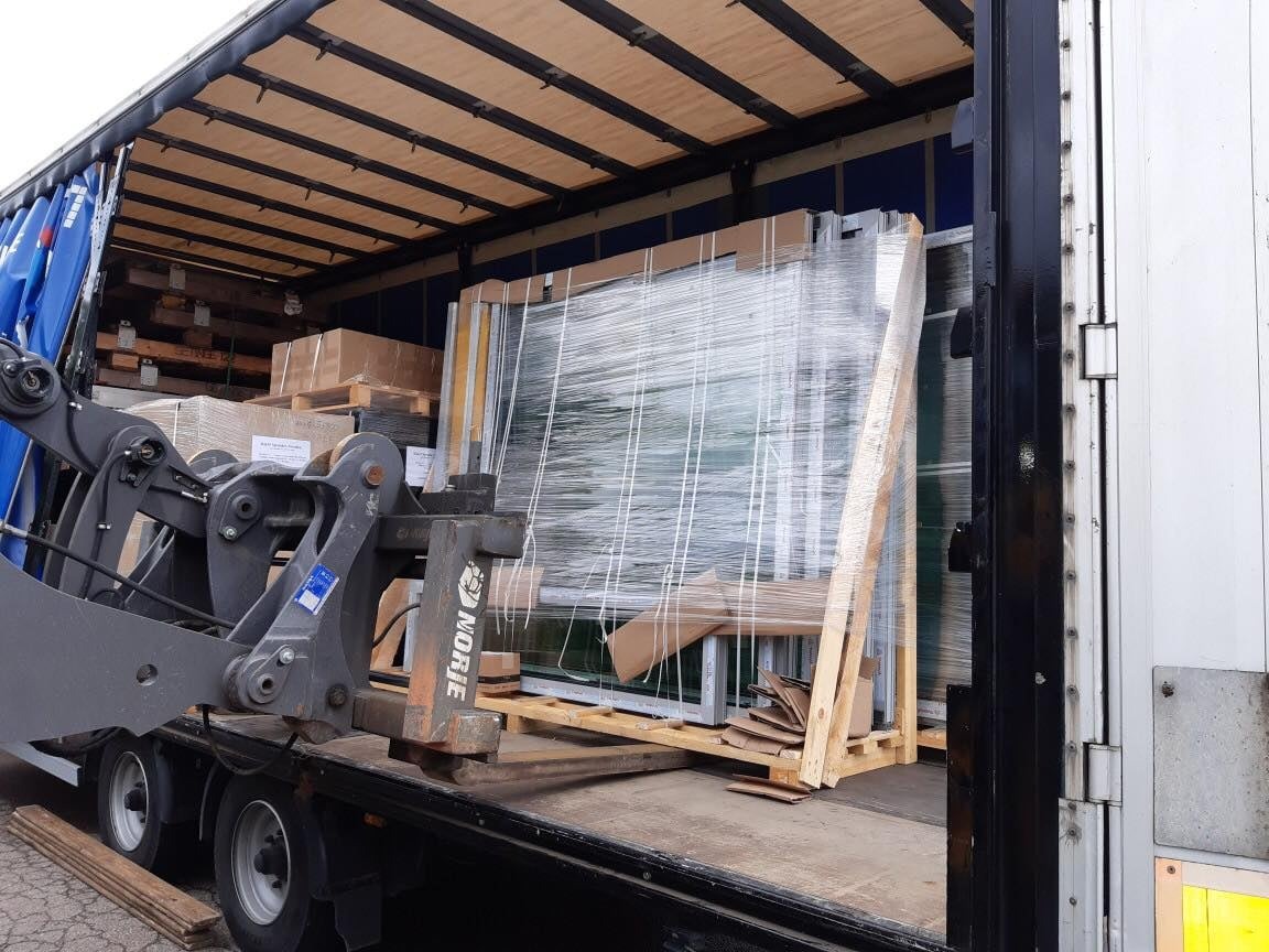 Wózek widłowy ładuje paletę z oknami owiniętymi folią do naczepy ciężarówki z niebieską plandeką, widoczne kartony i drewniane elementy zabezpieczające.