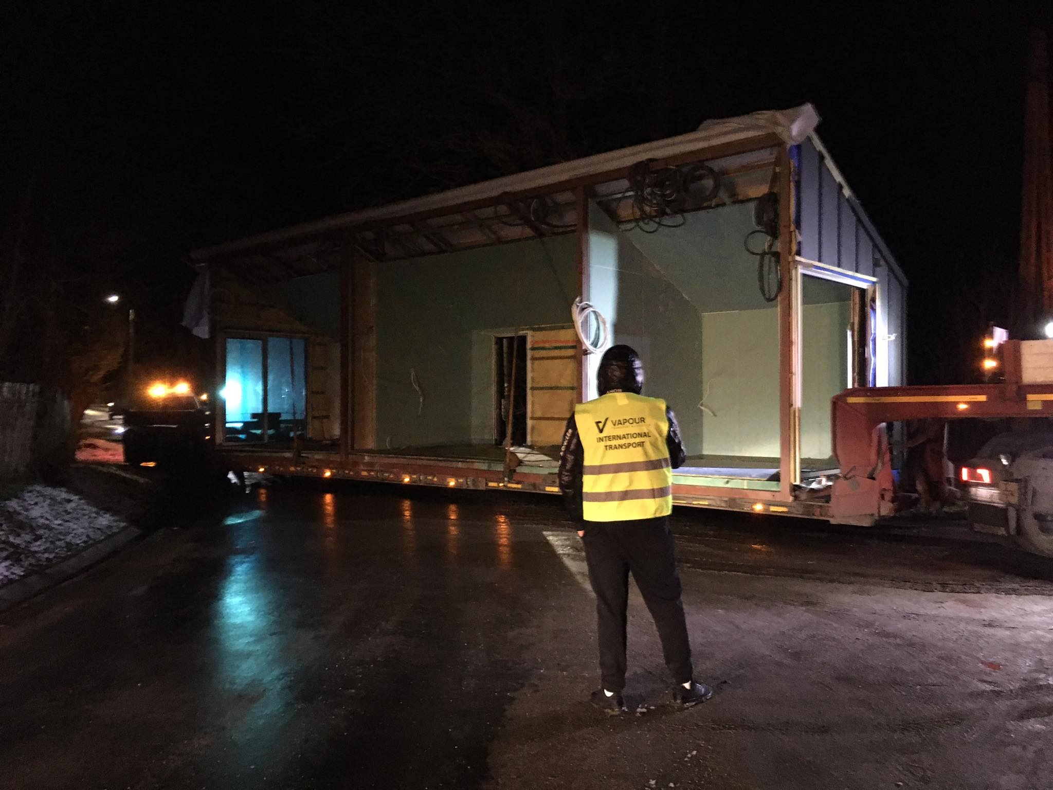 Nocny przewóz modułowego domu na lawecie, pracownik w kamizelce odblaskowej nadzoruje transport.