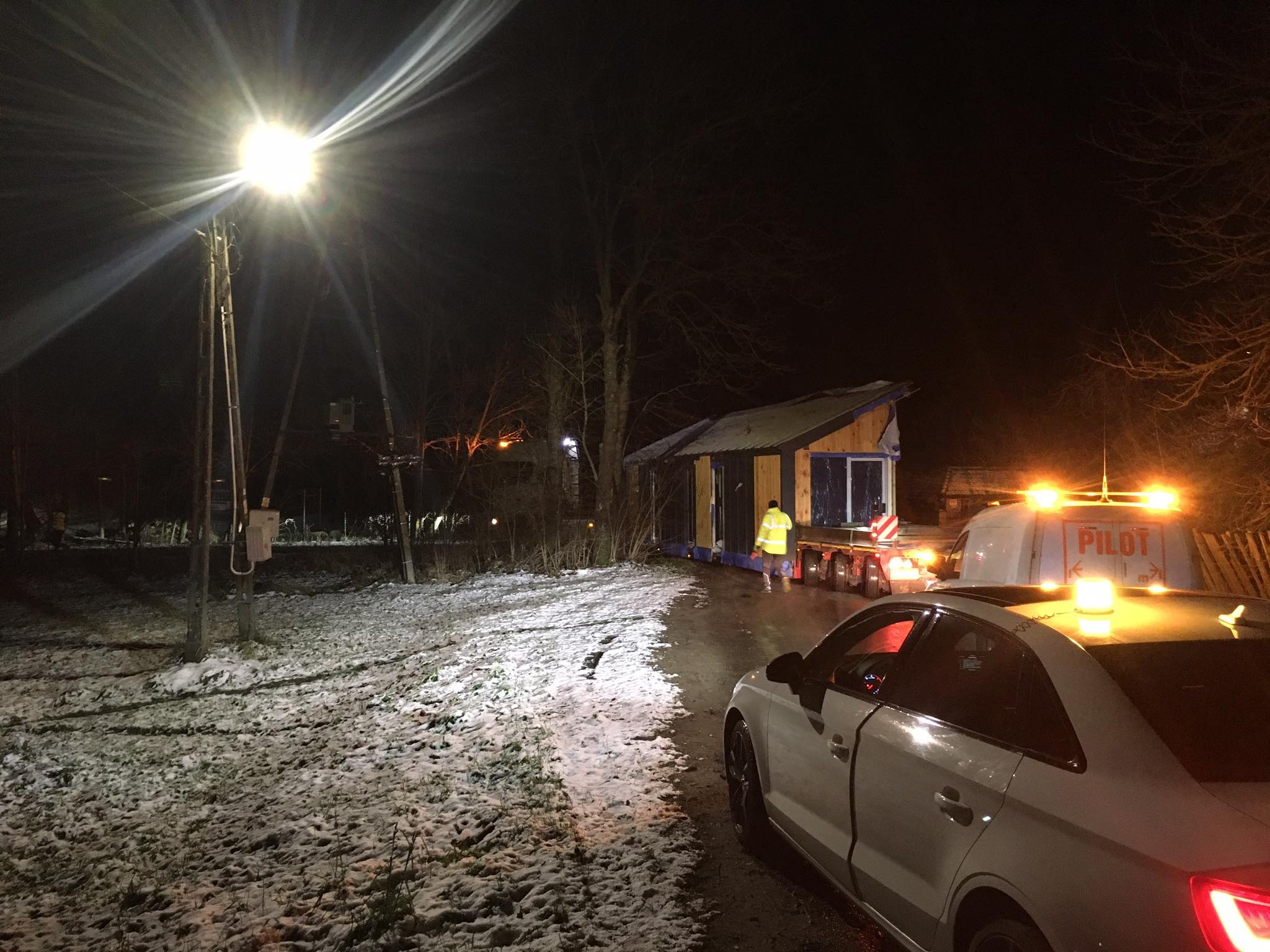 Nocny transport modułowego domu na platformie niskopodwoziowej, pilotowany przez pojazd z napisem 'PILOT', oświetlony latarnią uliczną; na pierwszym planie biały samochód osobowy.