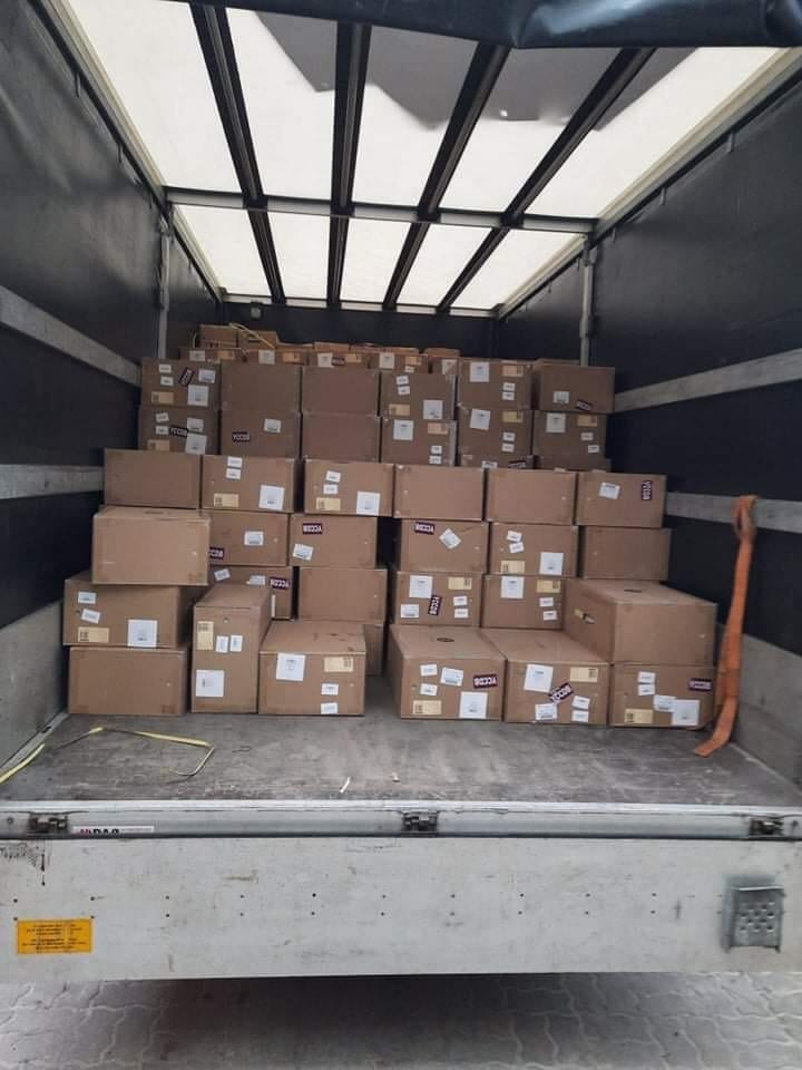 Wnętrze przestrzeni ładunkowej wypełnionej kartonami z etykietami, widoczne pasy zabezpieczające i pomarańczowy element mocujący przy ścianie, oświetlenie górne w przestrzeni ładunkowej.