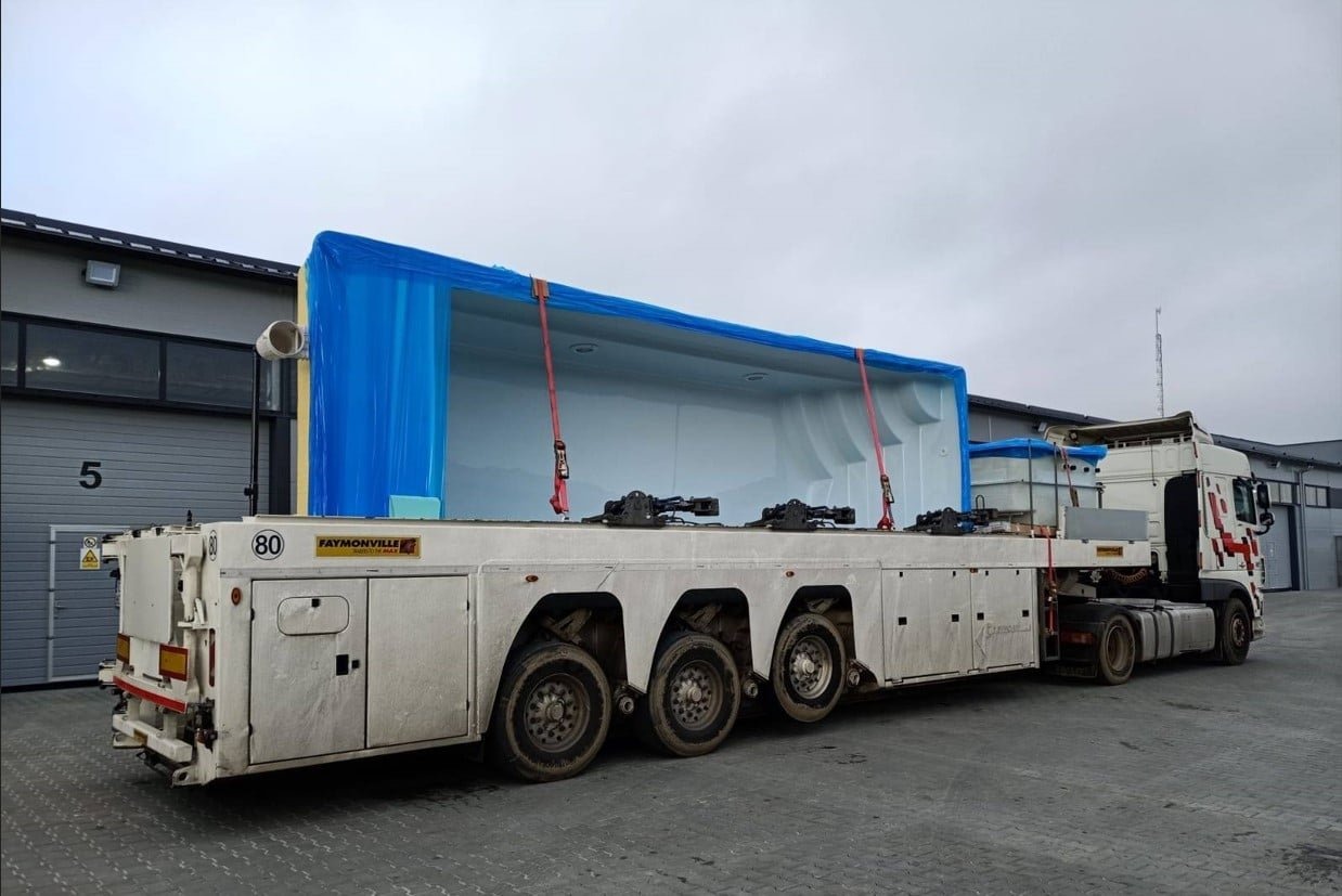 Nisko zawieszona naczepa Faymonville przewozi zabezpieczony niebieską folią duży, biały prefabrykat basenowy, widoczny numer 5 na budynku w tle.