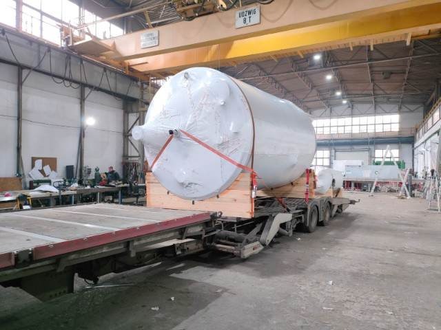 Duży, cylindryczny zbiornik owinięty folią, zabezpieczony czerwonymi pasami, umieszczony na drewnianej platformie na naczepie ciężarówki w hali przemysłowej z suwnicą stropową.