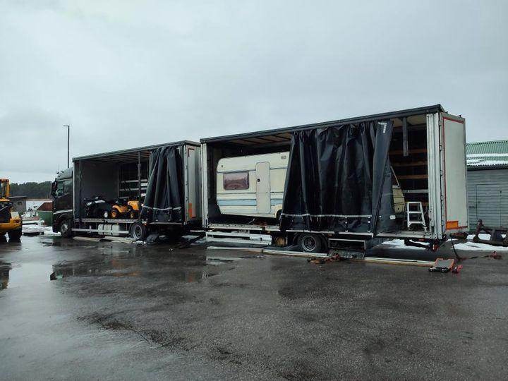 Dwa samochody ciężarowe z otwartymi naczepami, załadowane: jeden z przyczepą kempingową, drugi z dwoma traktorkami do koszenia trawy, na mokrym placu.