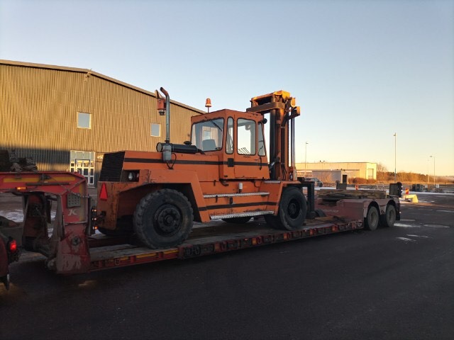 Pomarańczowy wózek widłowy na niskopodwoziowej przyczepie, widok z boku, na tle budynku przemysłowego i parkingu.