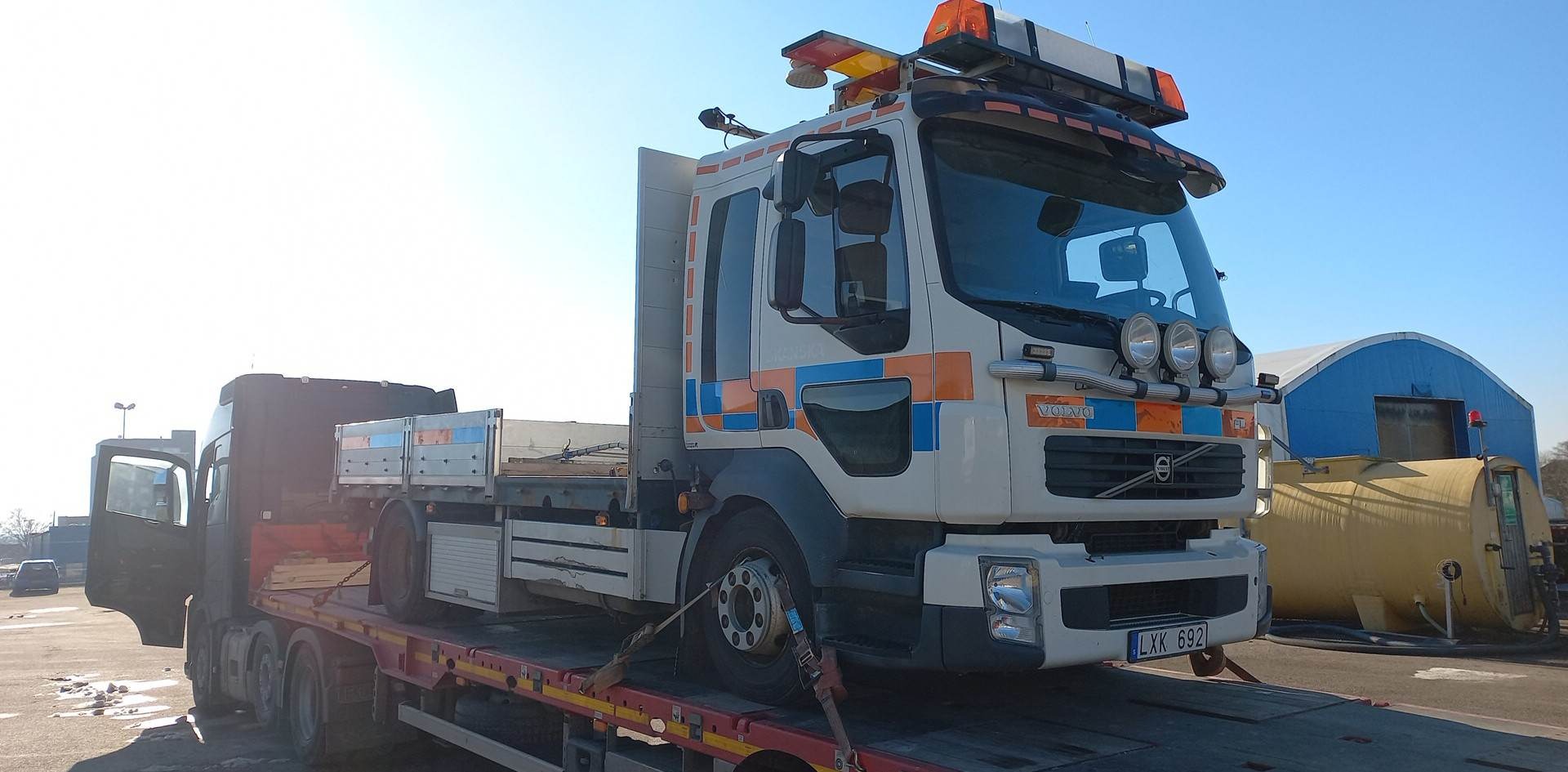 Ciężarówka Volvo z pomarańczowo-niebieskimi akcentami, z włączonymi światłami ostrzegawczymi na dachu, transportowana na niskopodwoziowej platformie, z widocznymi pasami mocującymi koła, na tle...