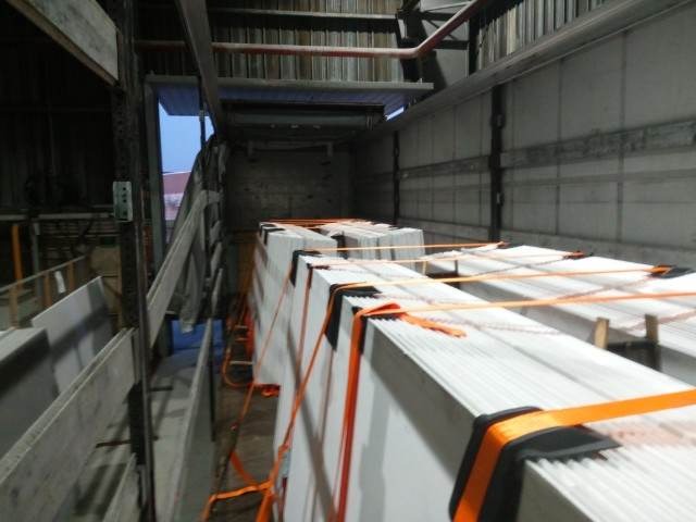 Wnętrze ciężarówki z ładunkiem płyt zabezpieczonych pomarańczowymi pasami transportowymi, widoczne metalowe elementy konstrukcyjne pojazdu.