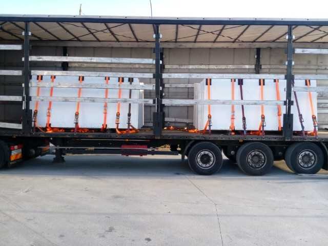 Ciężarówka z otwartą naczepą przewozi zabezpieczone pomarańczowymi pasami ładunki w białych opakowaniach.