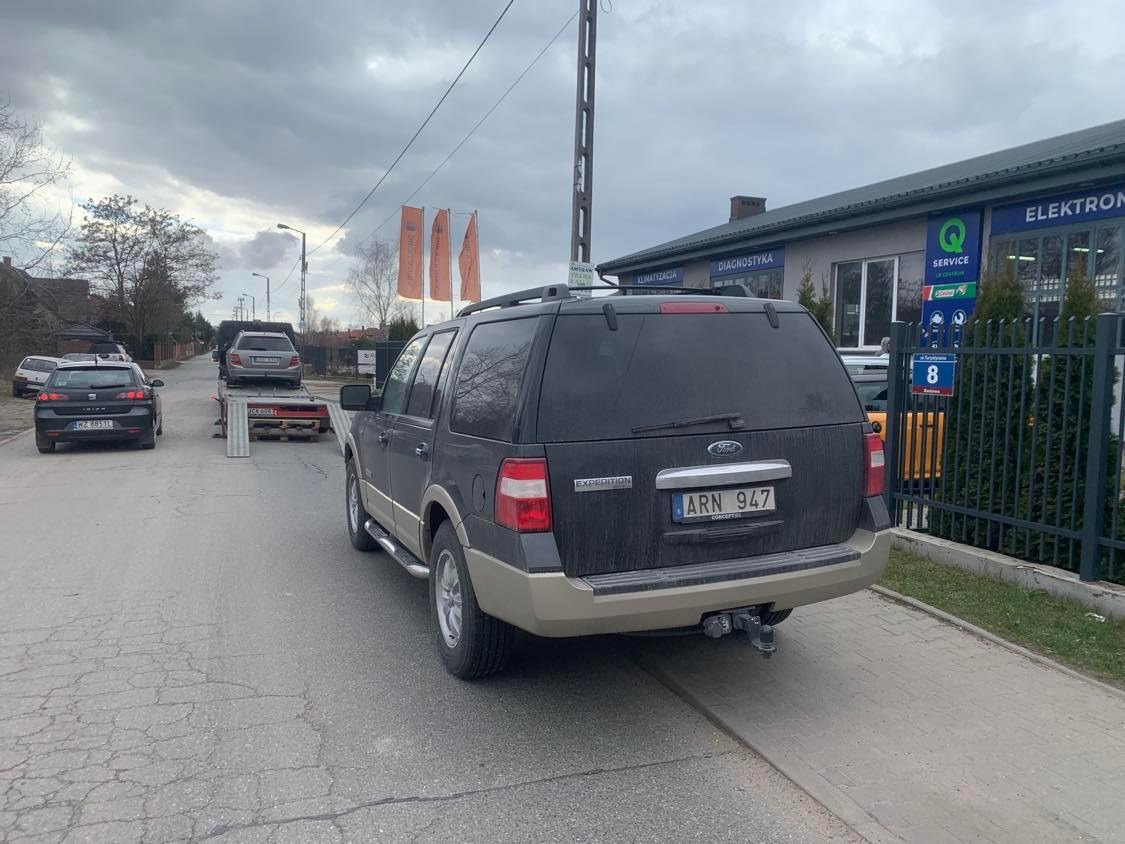Czarny Ford Expedition z przyczepą lawetą przewożącą srebrny samochód osobowy na tle zabudowań i pochmurnego nieba.