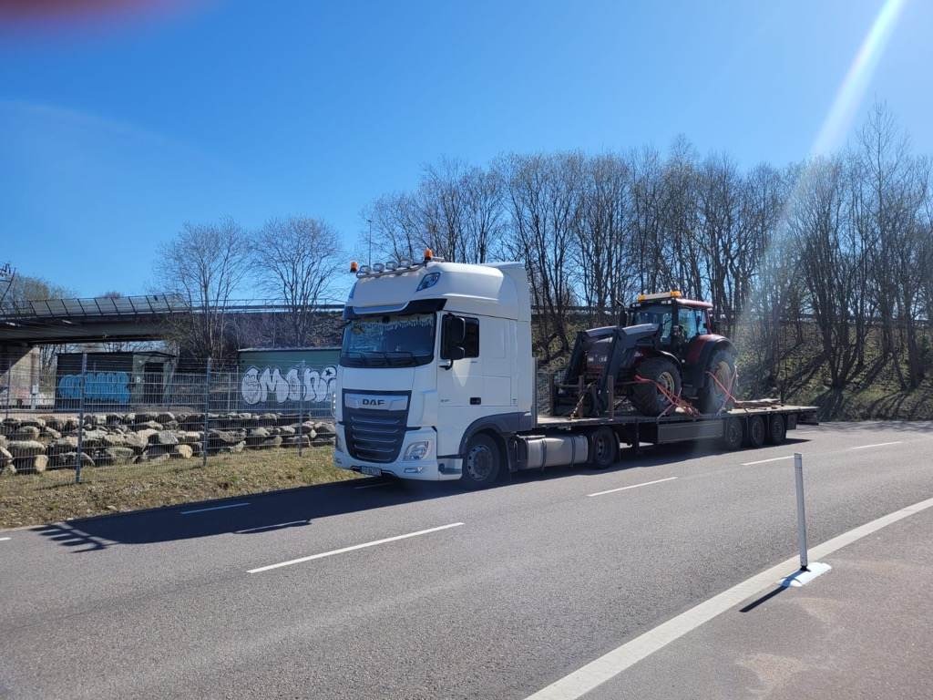 Ciężarówka DAF z naczepą przewozi czerwony traktor przez Warszawę; widoczny most w tle.