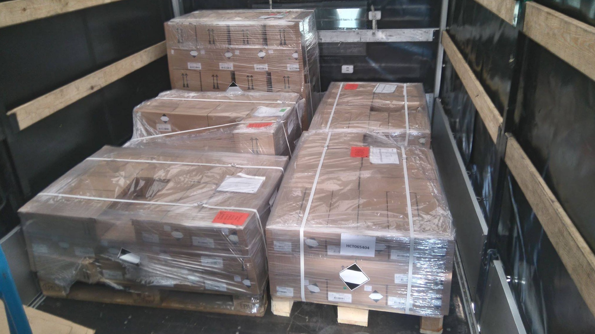 Kartonowe paczki zabezpieczone folią i pasami transportowymi, umieszczone na paletach w przestrzeni ładunkowej pojazdu, niektóre z oznaczeniami ostrzegawczymi.