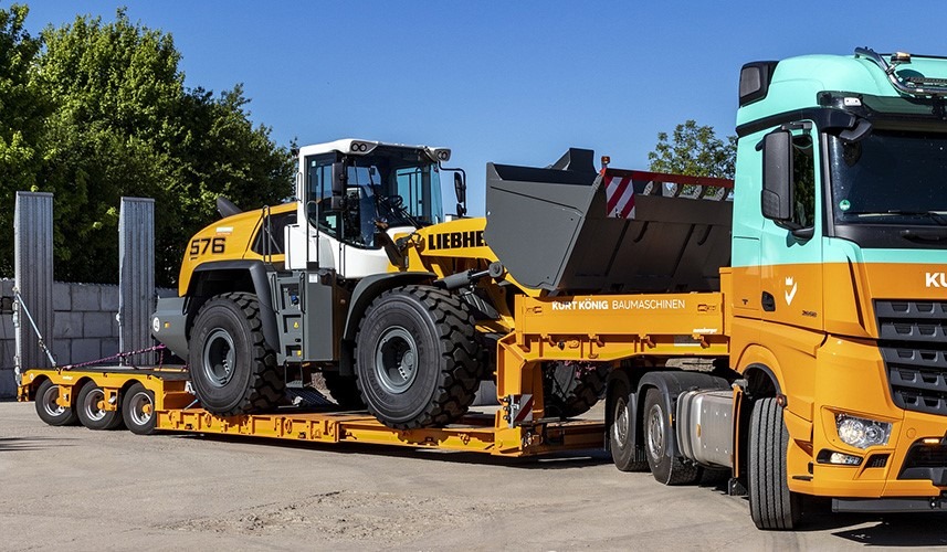 Żółto-szara ładowarka Liebherr 576 przewożona na niskopodwoziowej naczepie ciągniętej przez pomarańczowo-turkusowy ciągnik siodłowy, widok z boku na tle zieleni i błękitnego nieba.