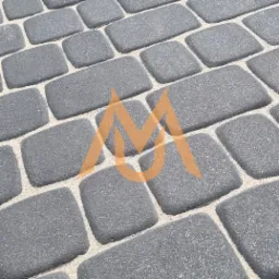 Szara kostka brukowa ułożona w regularny wzór, widok z bliska, z delikatnie zaokrąglonymi krawędziami i jasnymi fugami wypełnionymi piaskiem.