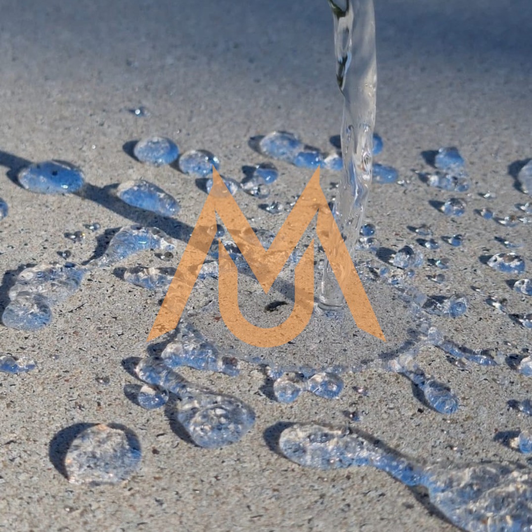 Strumień wody spływający po szarej, porowatej powierzchni betonowej, z widocznymi kroplami i plamami wody, nałożone logo w kolorze ochry.