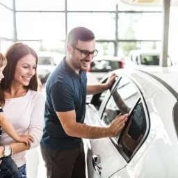 Rodzina z dzieckiem ogląda biały samochód w salonie dealera, mężczyzna sprawdza szybę, w tle inne pojazdy i duże okna.