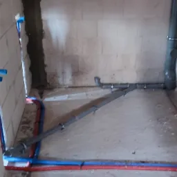 Surowy stan łazienki z widocznymi rurami wodnymi (czerwone i niebieskie) oraz kanalizacyjnymi (szare) ułożonymi na posadzce i przy ścianach z białego betonu komórkowego.