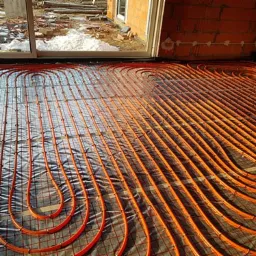 Pomarańczowe rurki ogrzewania podłogowego ułożone na srebrnej folii z siatką, widok z góry w surowym stanie, okno i mur z cegły w tle.