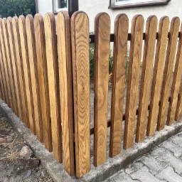 Fragment nowego, drewnianego ogrodzenia z zaokrąglonymi sztachetami, z widocznym fragmentem samochodu i chodnikiem z kostki.