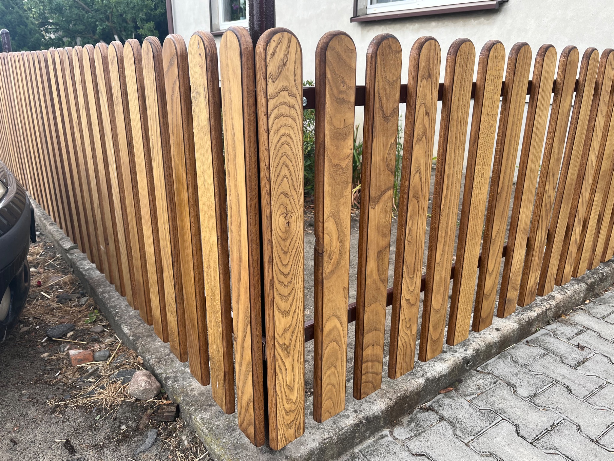 Fragment nowego, drewnianego ogrodzenia z zaokrąglonymi sztachetami, z widocznym fragmentem samochodu i chodnikiem z kostki.