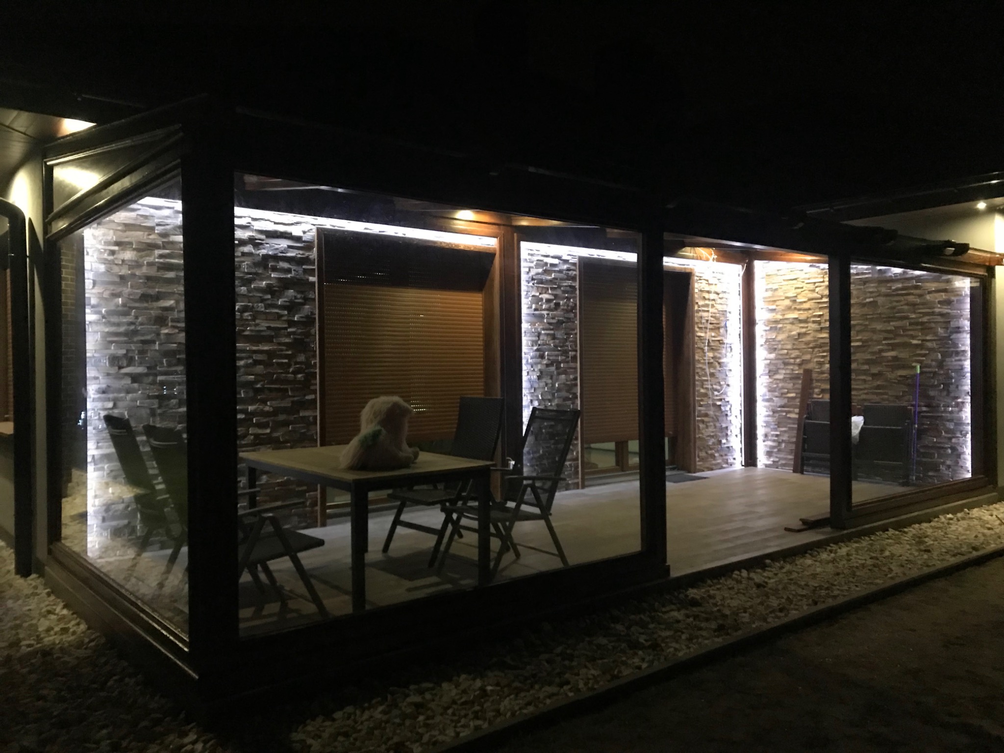 Zadaszony taras z kamienną ścianą podświetloną ledami, drewnianą podłogą i meblami ogrodowymi widziany nocą z zewnątrz.