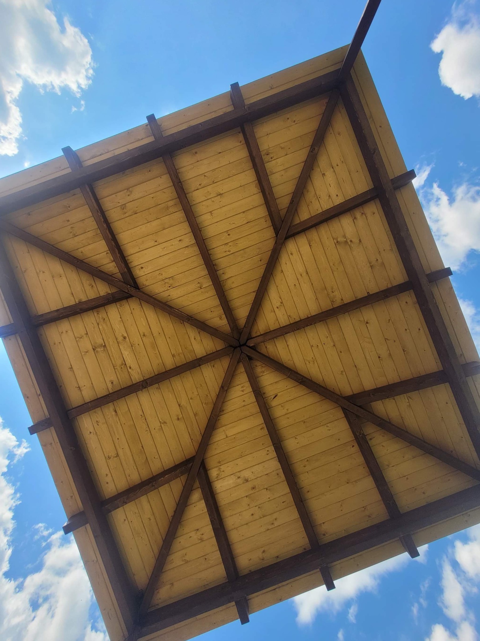 Widok z dołu na drewniany taras w Noskowie, z jasnymi deskami i ciemnymi belkami na tle błękitnego nieba z chmurami.