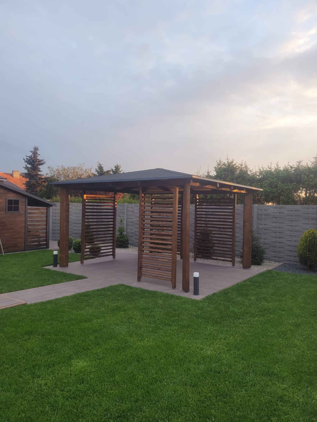 Drewniana altana z ażurowymi ścianami na tarasie z kostki brukowej, otoczona trawnikiem i ogrodzeniem panelowym w ogrodzie.