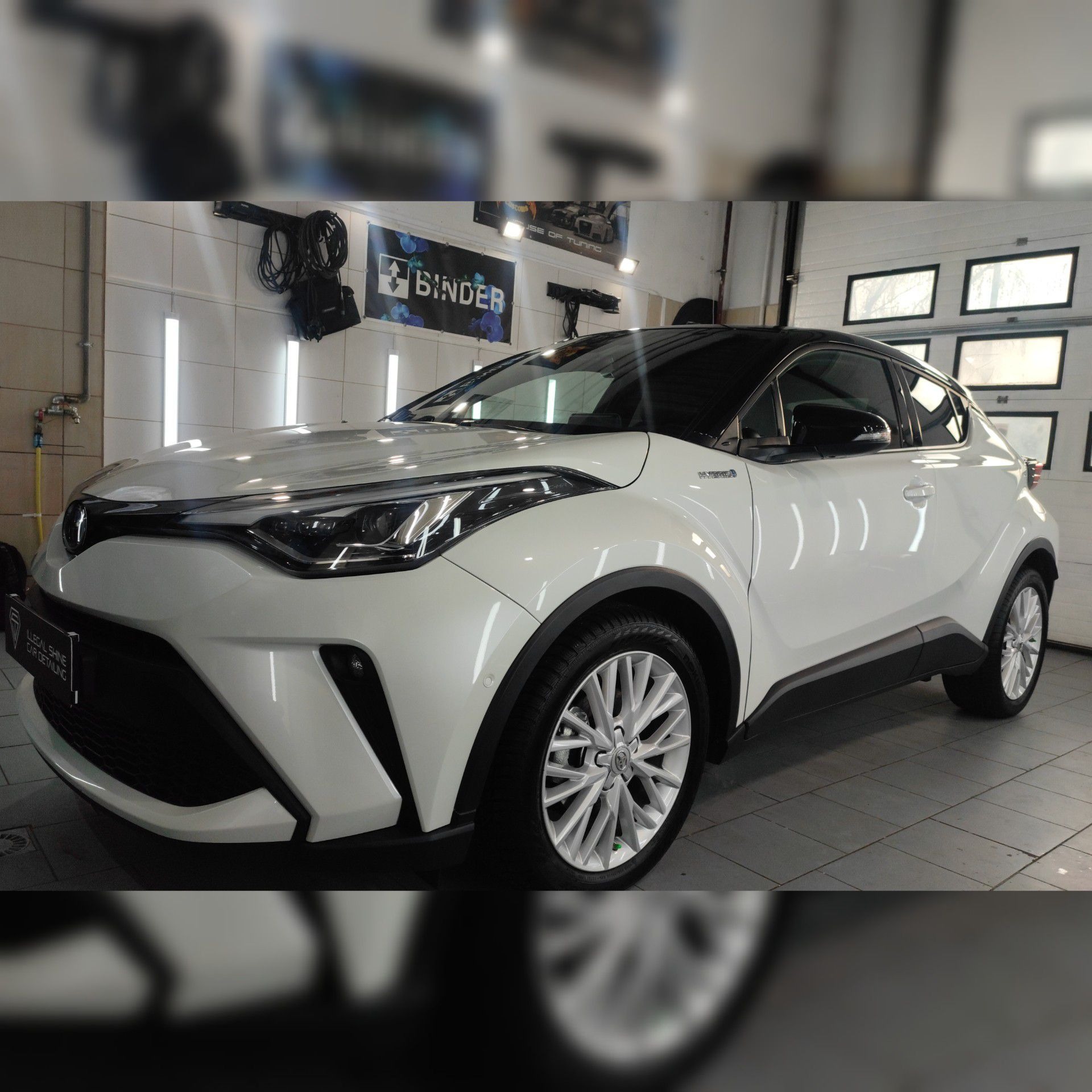 Biały samochód Toyota C-HR po umyciu, widoczne krople wody na lakierze, na tle myjni z oświetleniem ledowym i banerem reklamowym.