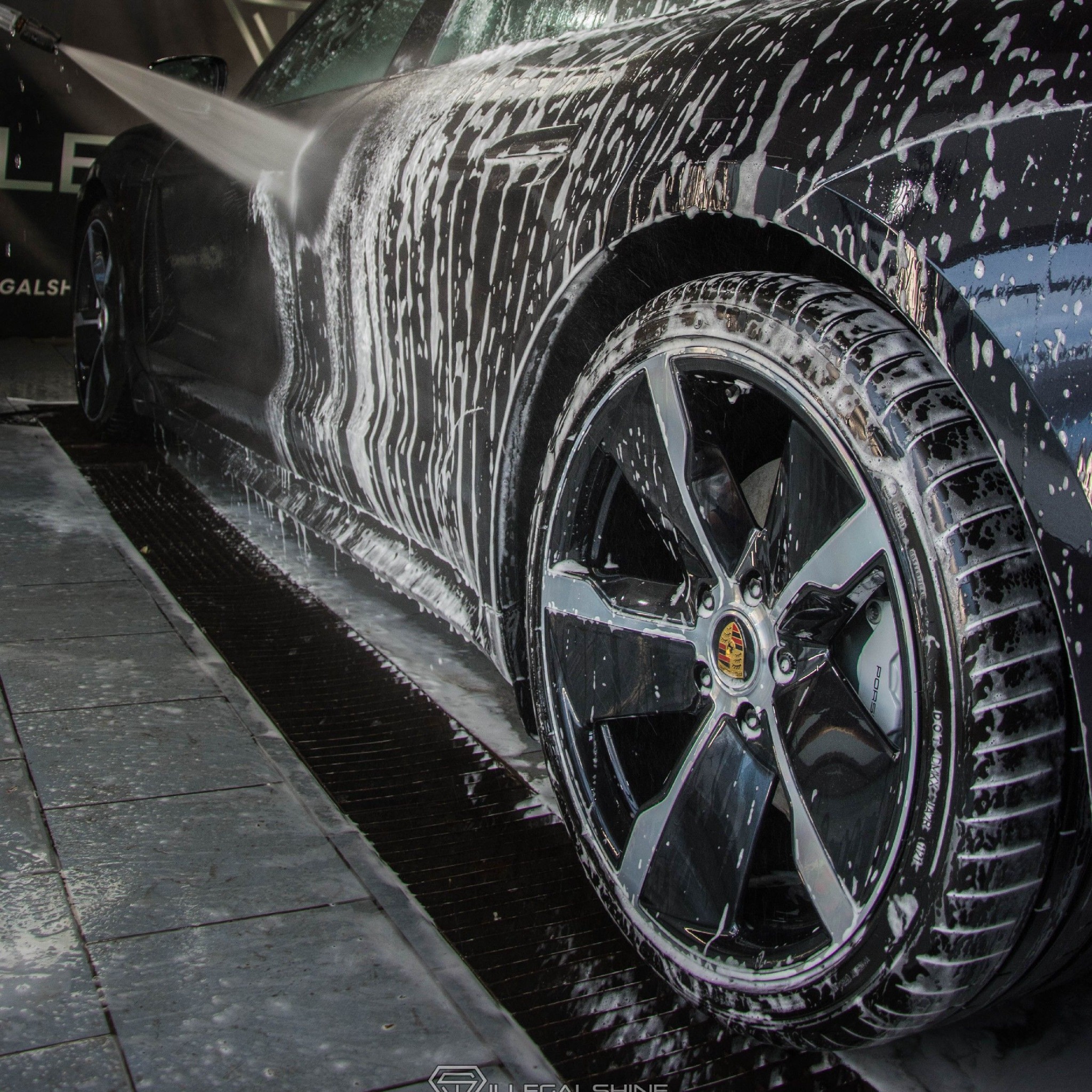Czarny samochód Porsche pod ciśnieniem wody z pianą, widoczne felgi i opony z bliska, w tle logo firmy.