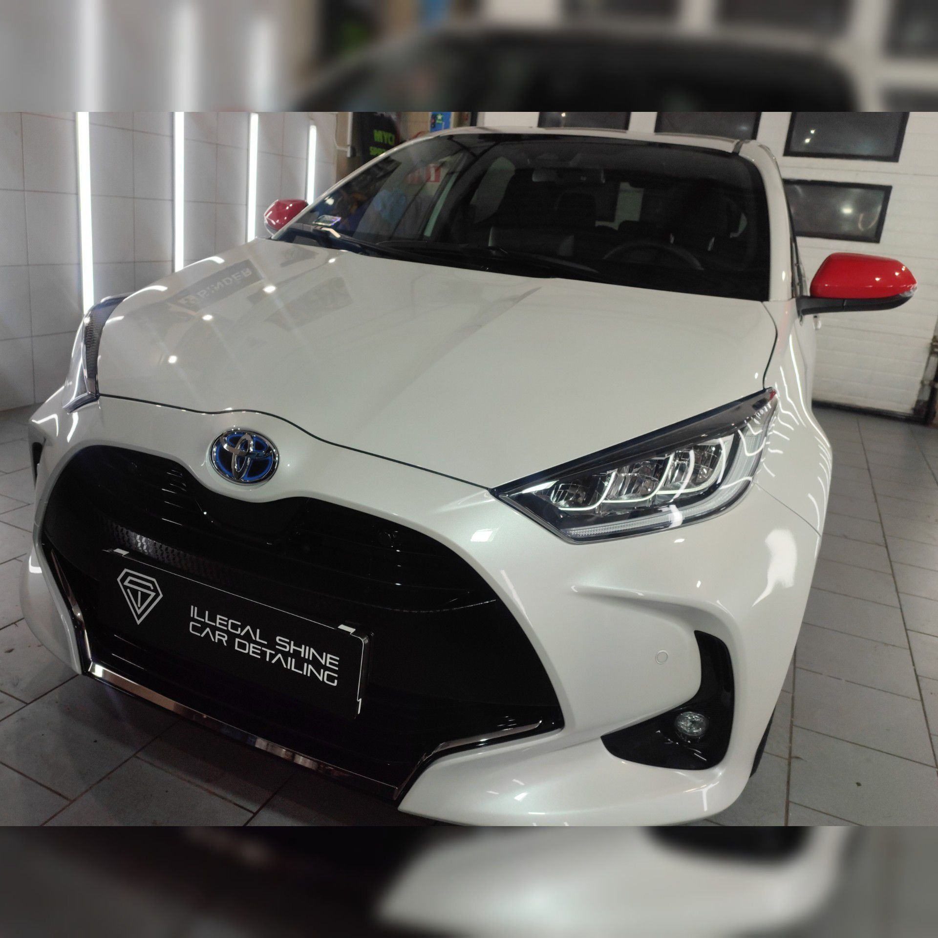 Biała Toyota Yaris po detailingu z czerwonymi lusterkami, widoczne logo firmy Illegal Shine Car Detailing na czarnym panelu.
