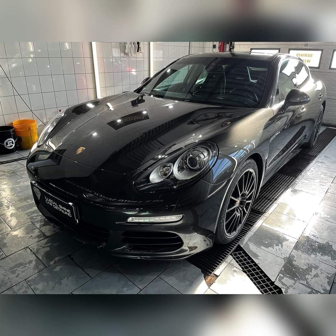 Błyszczący, czarny Porsche Panamera po usłudze detailingu, widoczne odbicia światła w lakierze, logo firmy detailingowej na przednim zderzaku.
