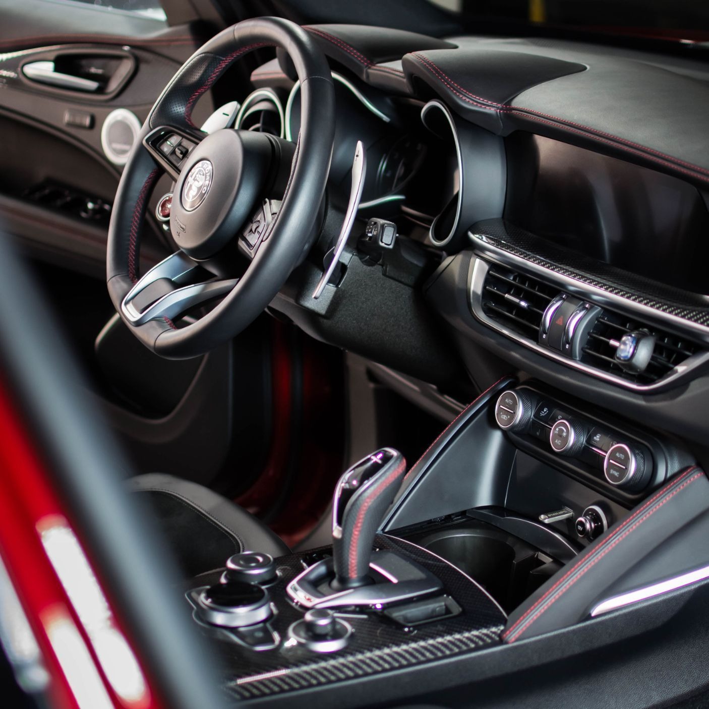 Wnętrze samochodu Alfa Romeo z czarną skórzaną tapicerką przeszytą czerwoną nicią, widok na deskę rozdzielczą, kierownicę z logo i konsolę środkową.