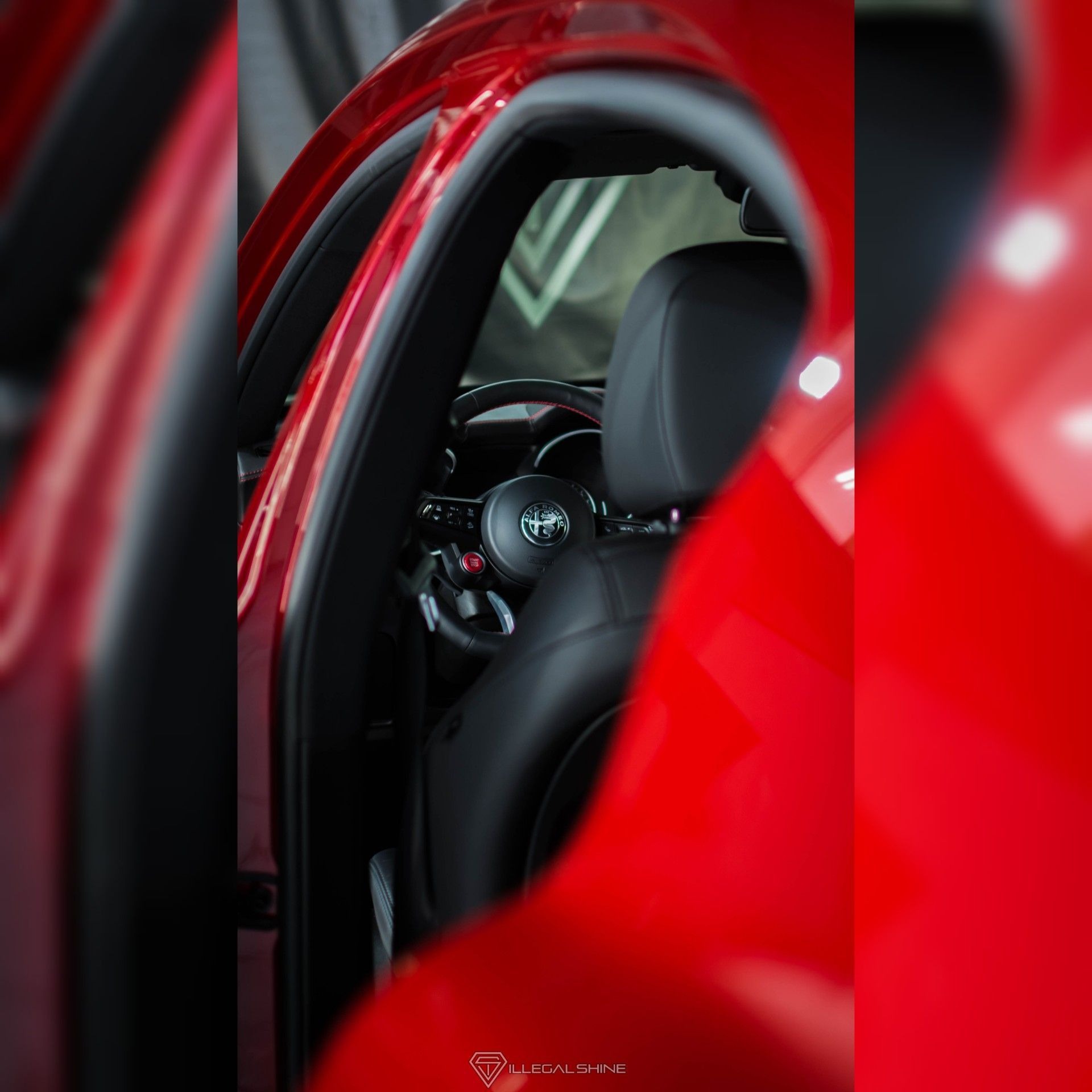 Wnętrze czerwonego samochodu Alfa Romeo, widok z zewnątrz przez otwarte drzwi na czarną skórzaną tapicerkę fotela i kierownicę z logo marki.