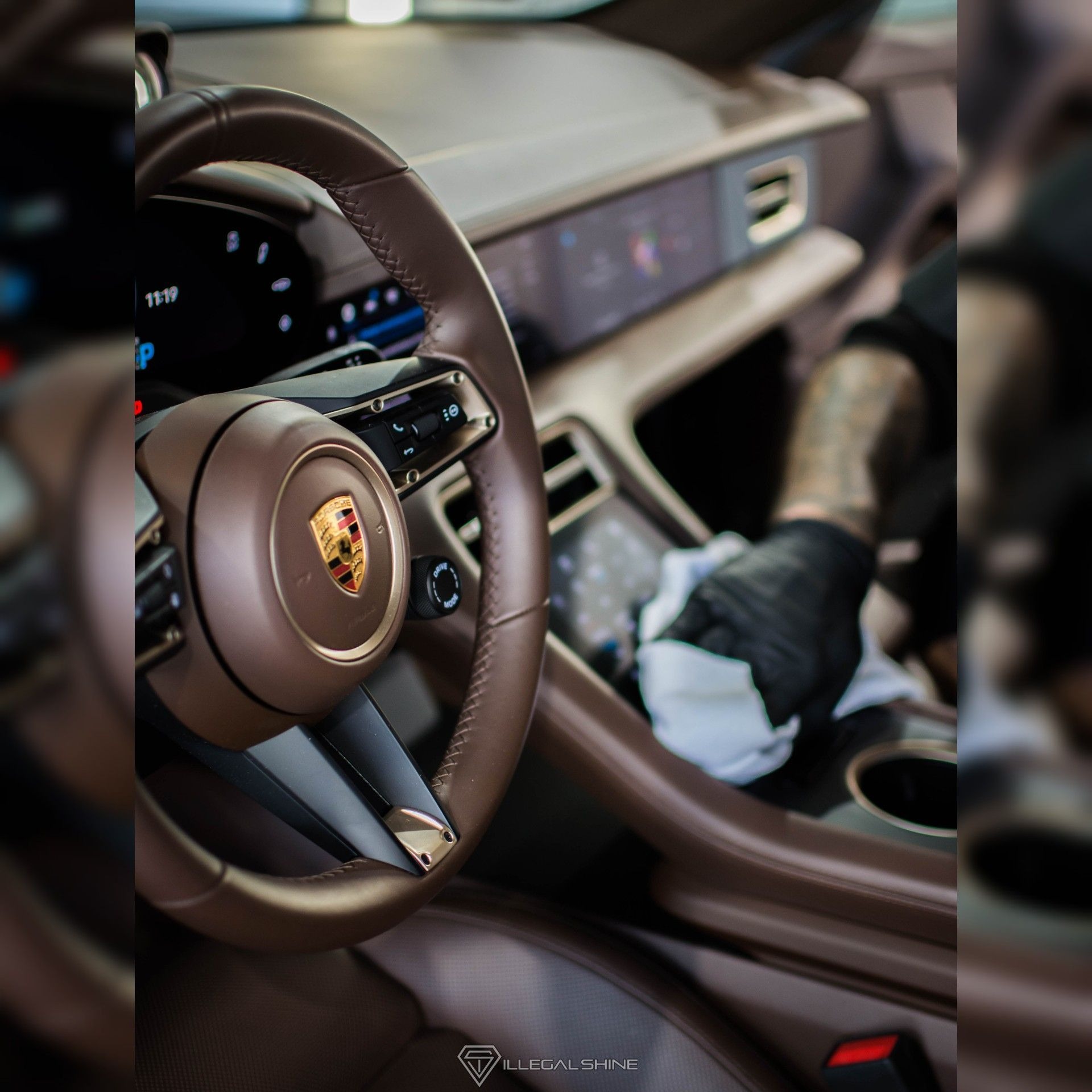 Wnętrze Porsche Taycan czyszczone przez osobę w czarnych rękawiczkach, widoczny brązowy skórzany kokpit i kierownica z logo producenta.