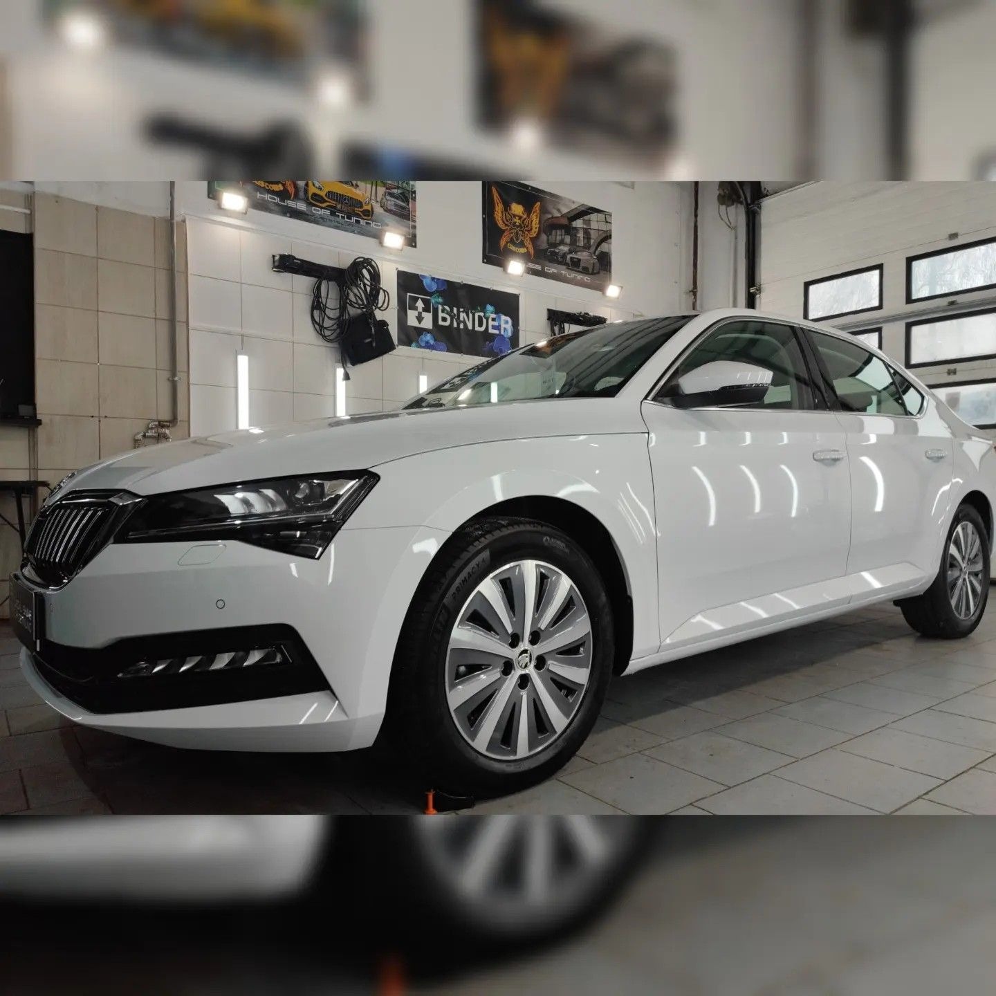Biała Skoda Superb po umyciu w myjni, widoczne odbicia światła na lakierze, w tle plakat z motywem tuningowym i akcesoria do mycia.