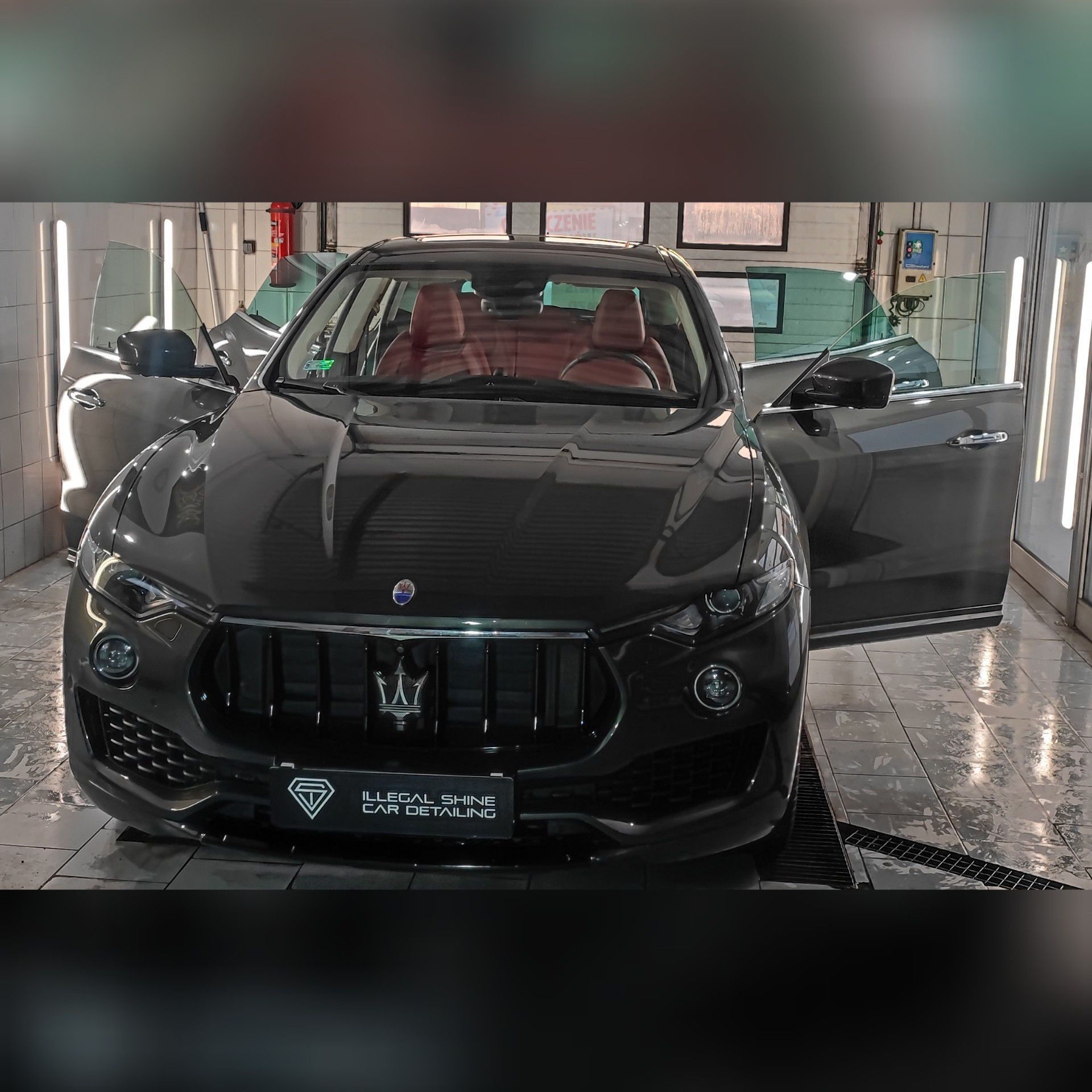 Szary Maserati Levante z otwartymi drzwiami, widoczne czerwone skórzane fotele wewnątrz, stoi w jasnej hali myjni z kratką odpływową w podłodze; logo firmy detailingowej na przednim zderzaku.