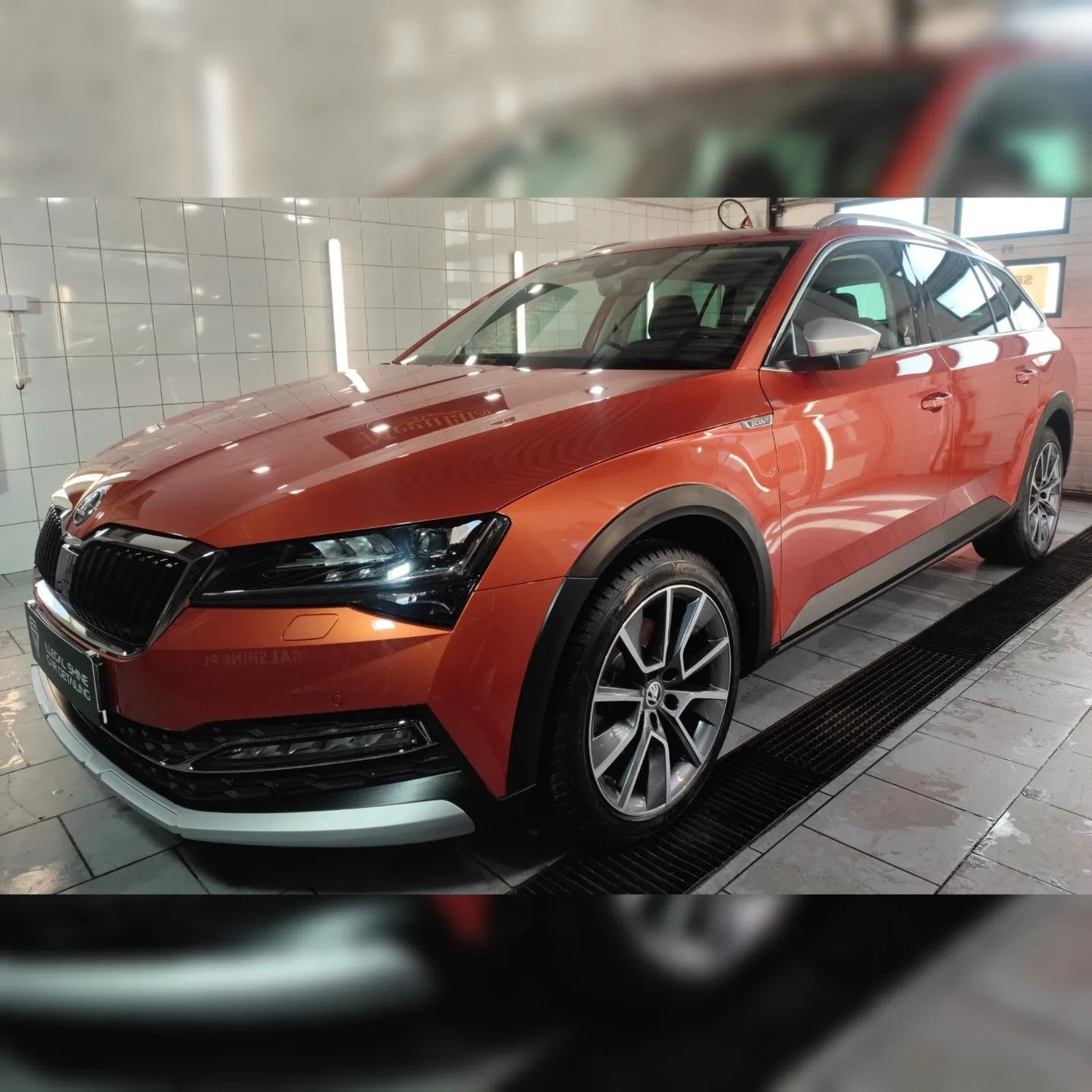 Pomarańczowy samochód Skoda Superb Scout po umyciu w myjni, widoczny z przodu pod kątem, z połyskiem na karoserii i mokrą podłogą w tle.
