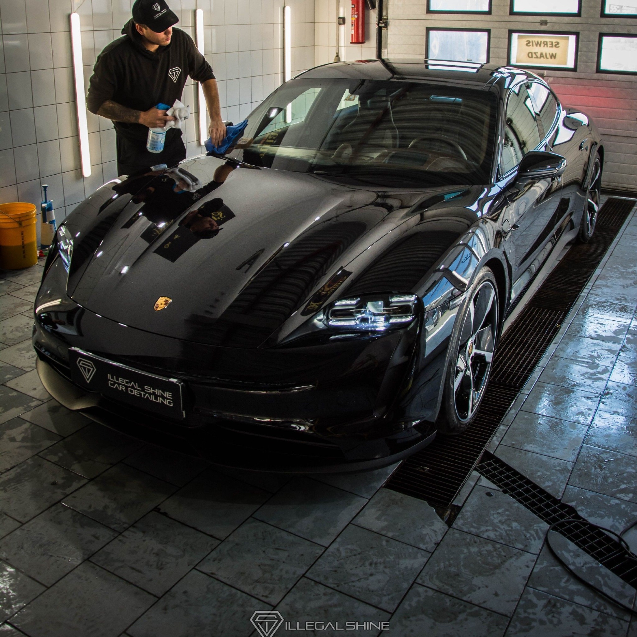 Pracownik w czarnej bluzie z kapturem i czapce czyści czarnego Porsche Taycan w myjni, używając spryskiwacza i niebieskiej ściereczki z mikrofibry, widoczny napis 'ILLEGAL SHINE CAR DETAILING'...