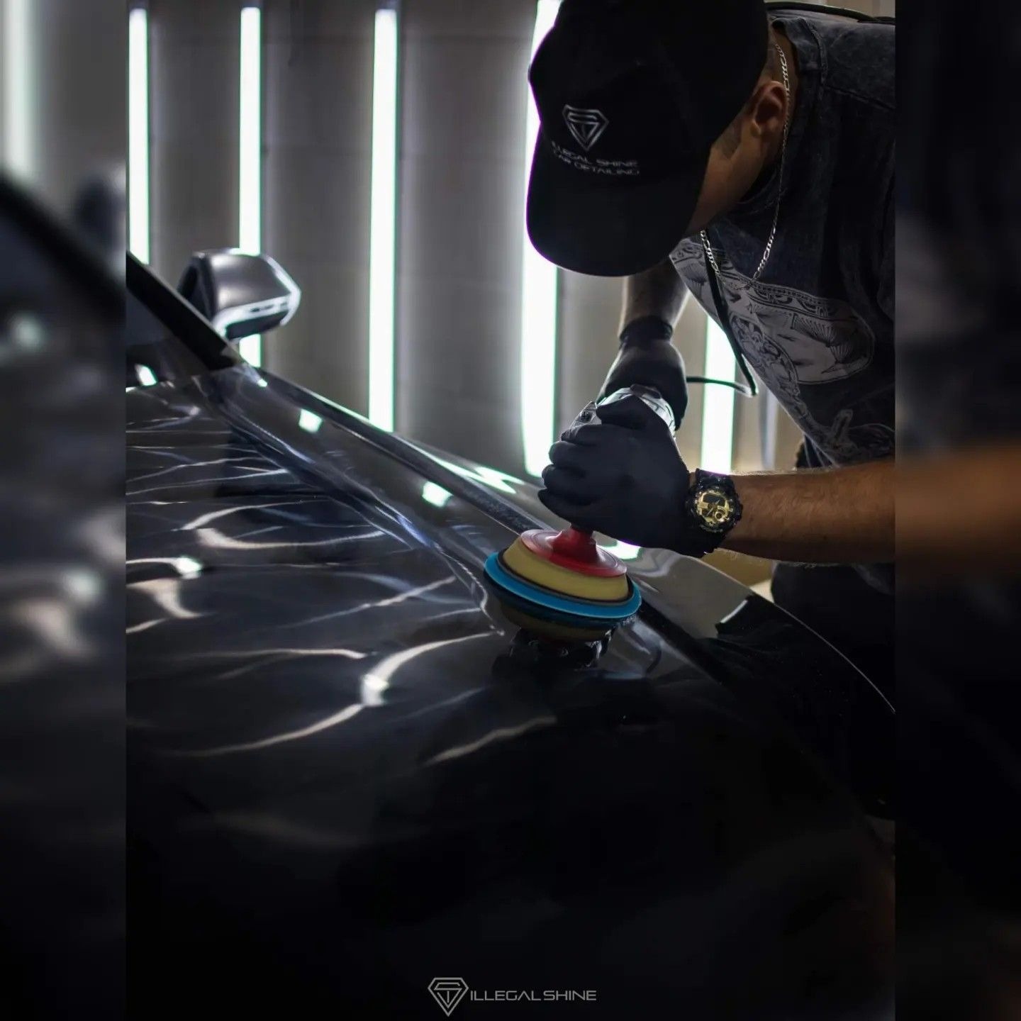 Polerowanie czarnego lakieru samochodowego za pomocą polerki rotacyjnej z kolorowymi padami, widoczny operator w czarnych rękawicach i czapce z logo firmy, oświetlenie warsztatowe w tle.